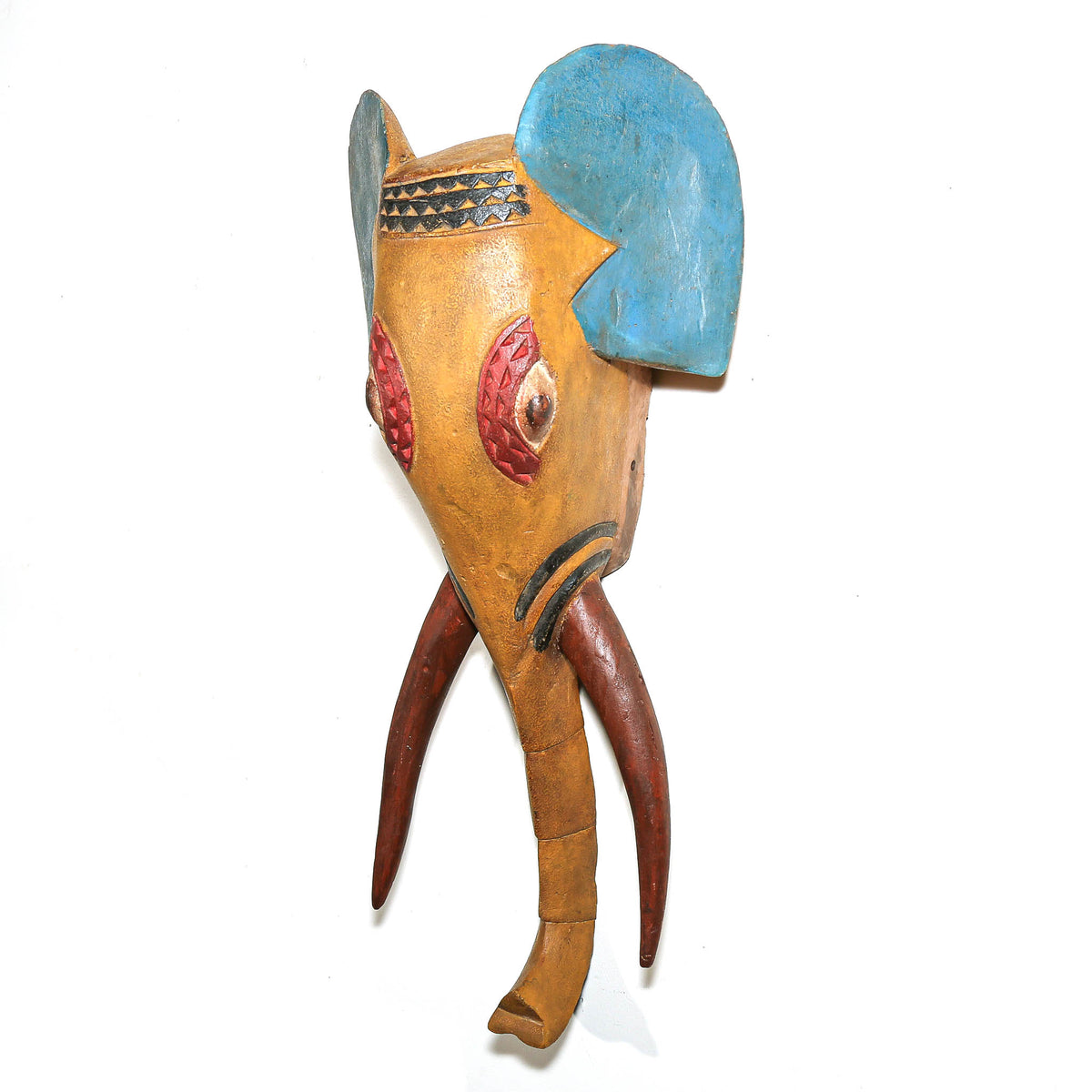 A handcrafted wooden elephant mask with a natural tan body, a bright blue ear, reddish-pink eye details, and brown tusks, shown against a plain white background.