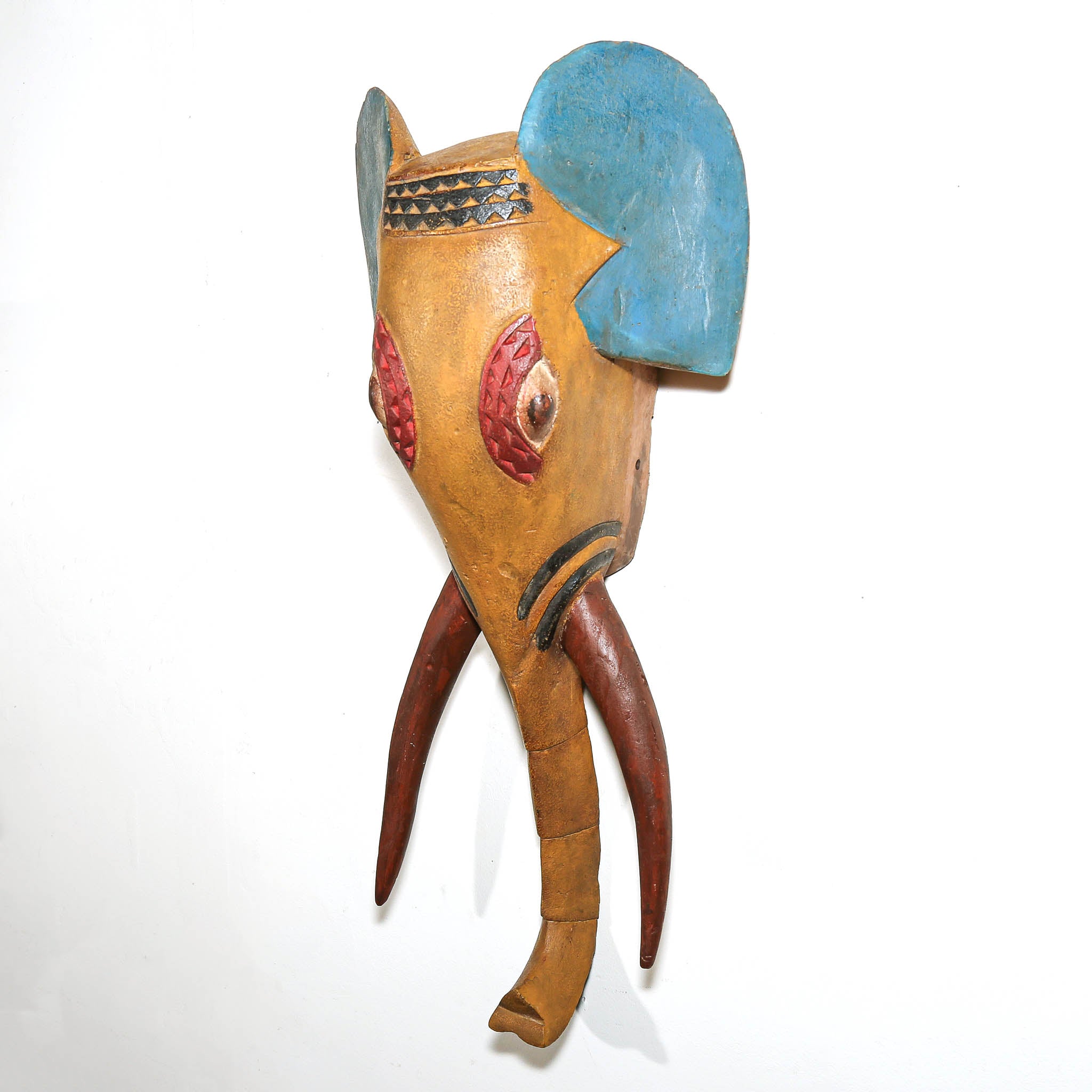 A hand-carved African wooden elephant mask with a natural tan wood body, painted blue ears, red patterned eyes, and dark brown tusks, shown against a plain white background.