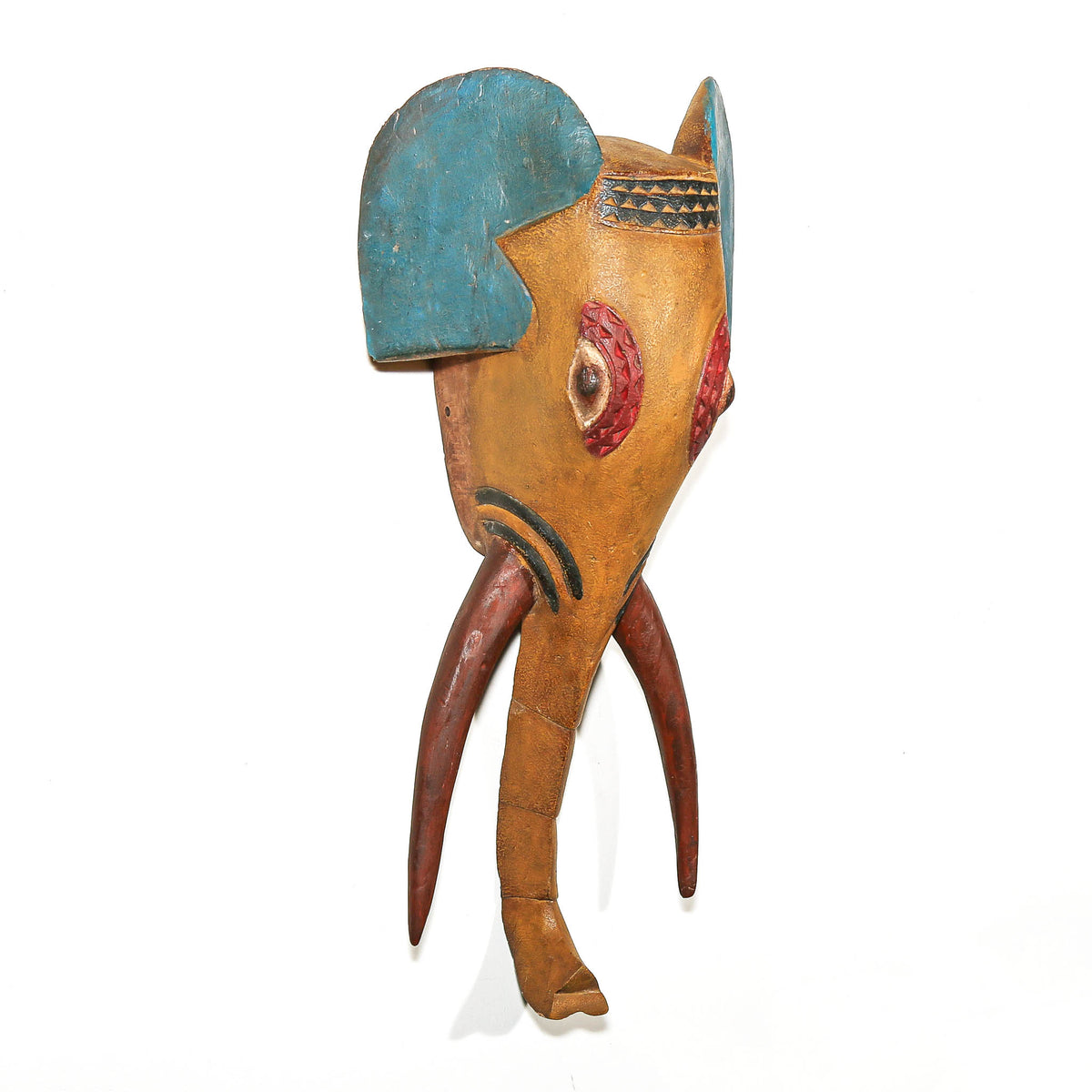A hand-carved wooden African-style elephant mask with a natural tan wood body, painted blue ear, red eye, and reddish-brown tusks, shown against a plain white background.