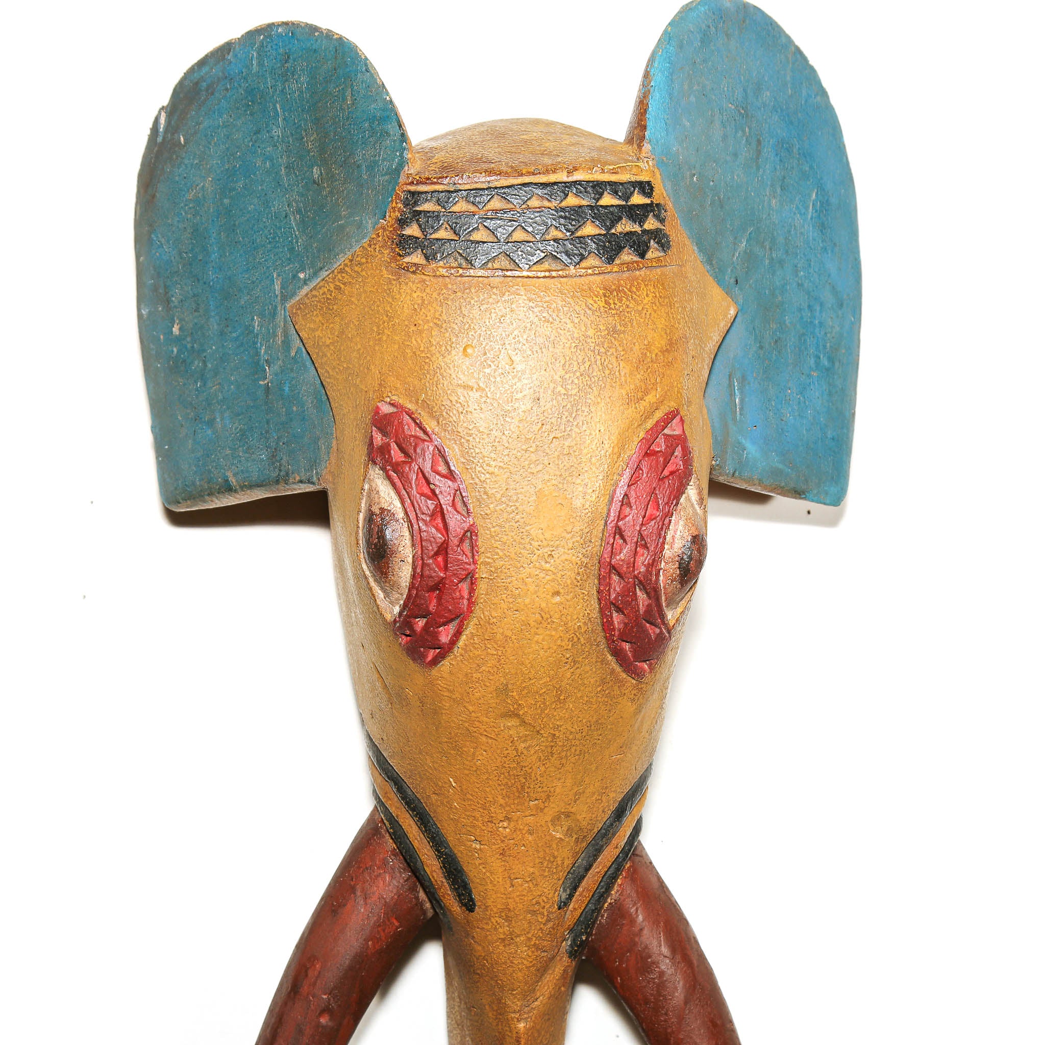 A handcrafted wooden elephant head figurine with a weathered tan-brown body, large blue ears, red patterned eyes, and black decorative details, shown against a plain white background.