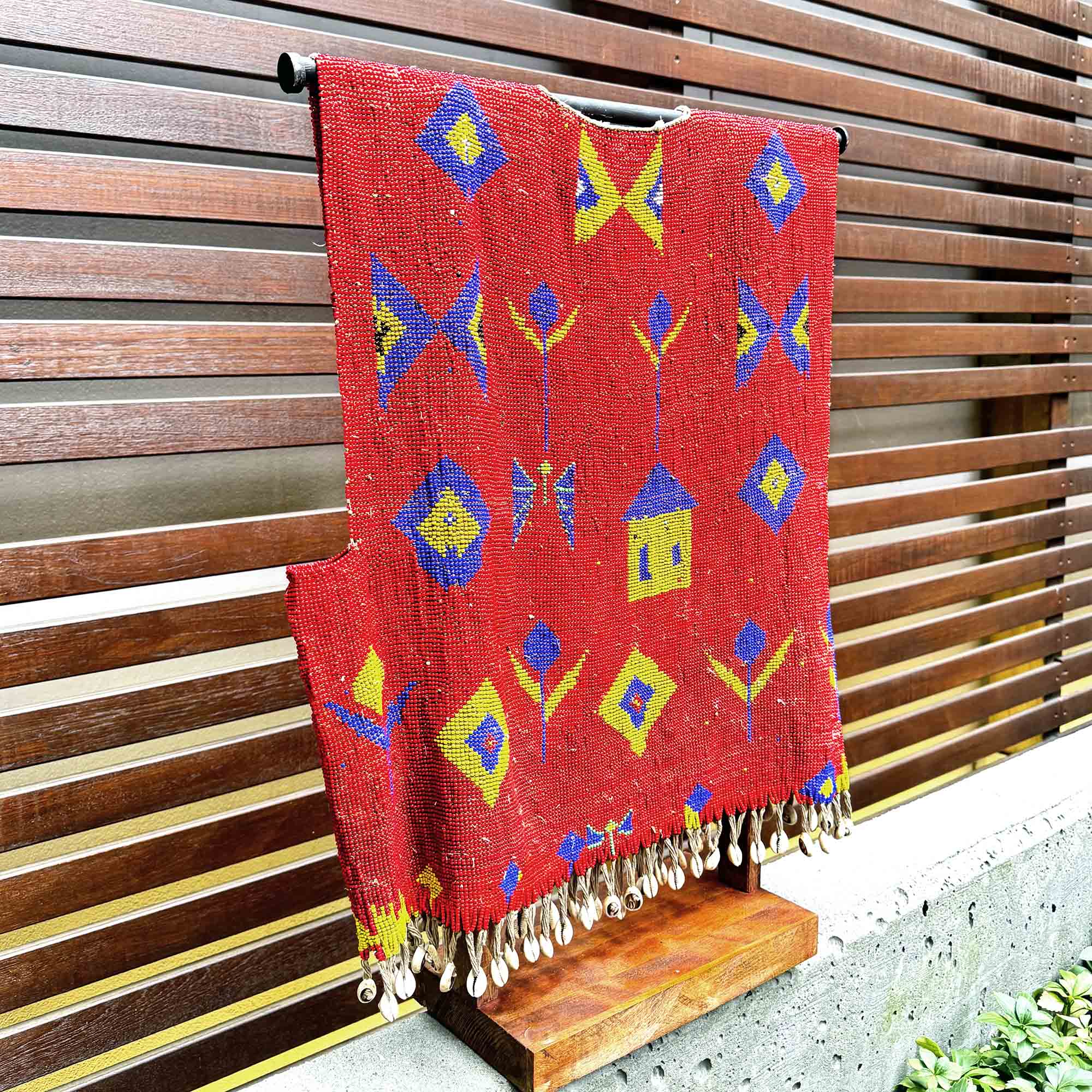 Red woven beaded tunic with colorful patterns hanging on a wooden stand.