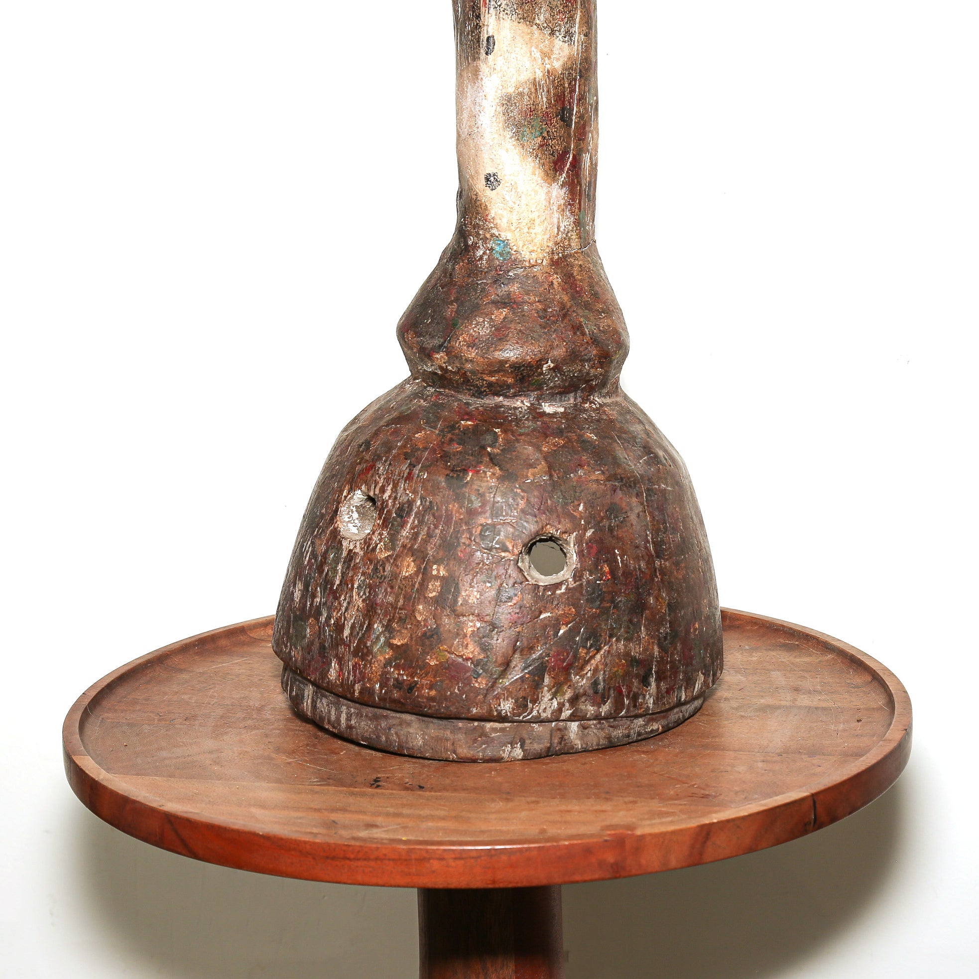 A rustic, hand-carved wooden sculpture with mask eye holes or decorative table piece with a textured, weathered brown and beige finish, featuring a circular wooden base and a tall, narrow neck, shown against a plain white background.