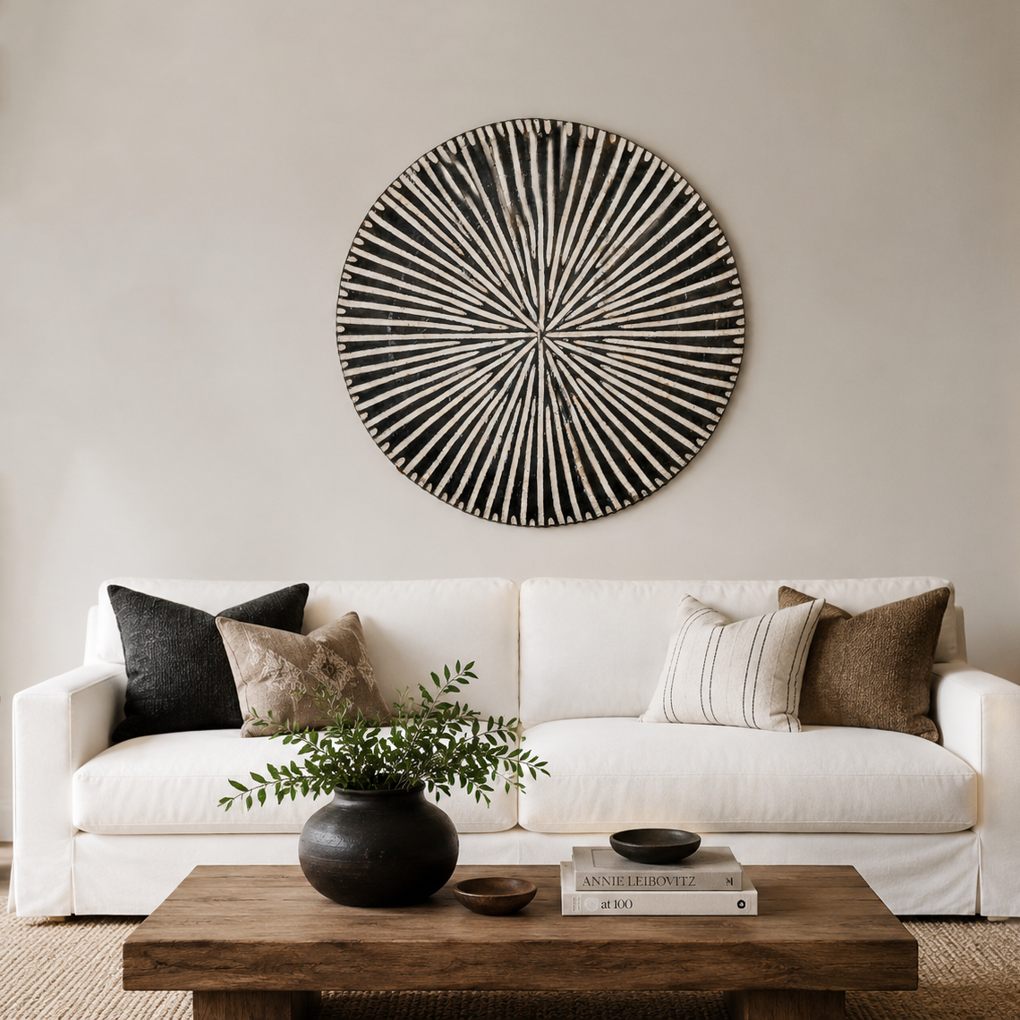 A styled living room scene featuring a white fabric sofa with black, beige, and brown throw pillows, a rustic wooden coffee table with a black vase and books, and a large circular black and white woven wall art piece mounted on the beige wall above the sofa.