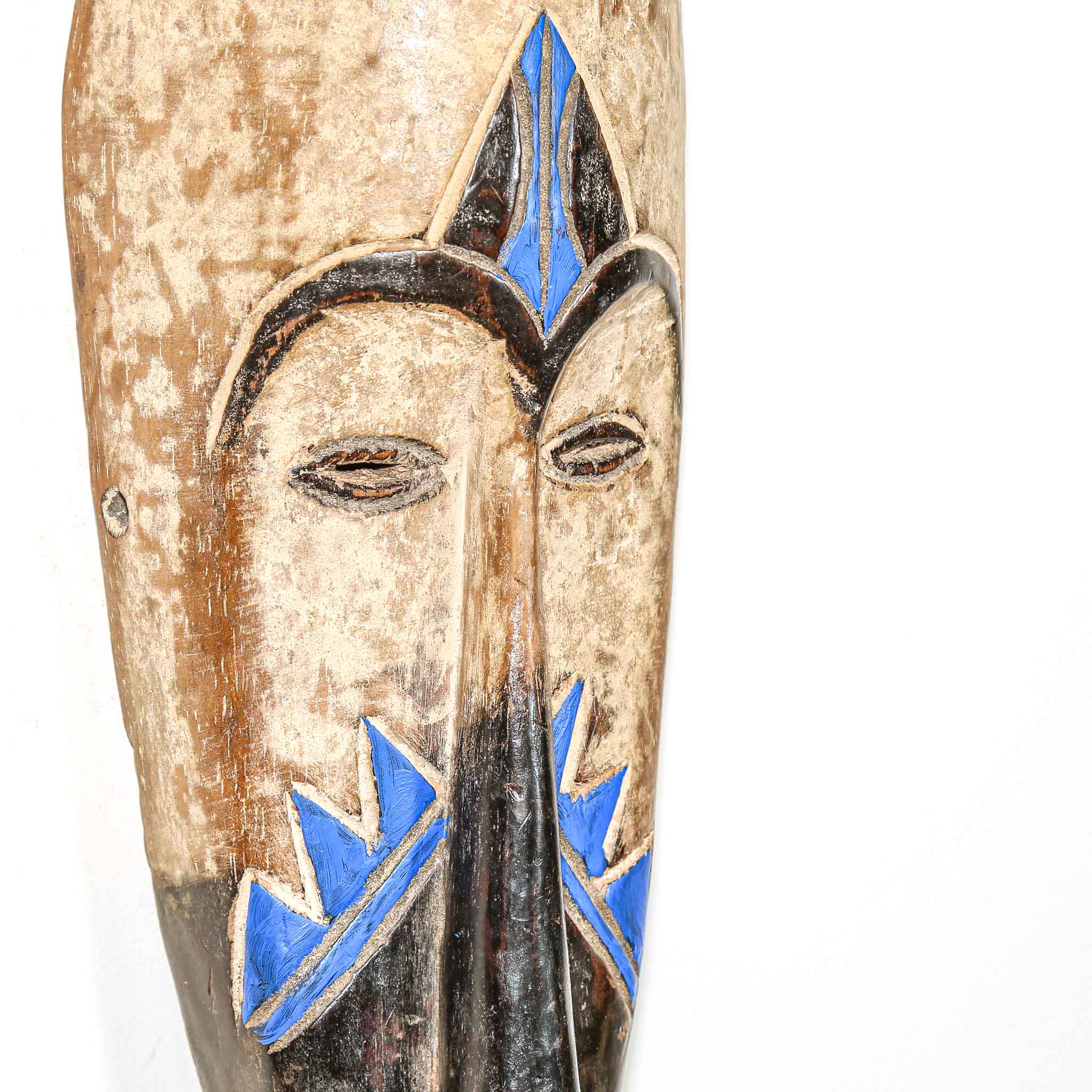 Close up of a sculpted wooden mask with blue accents on a white background wall