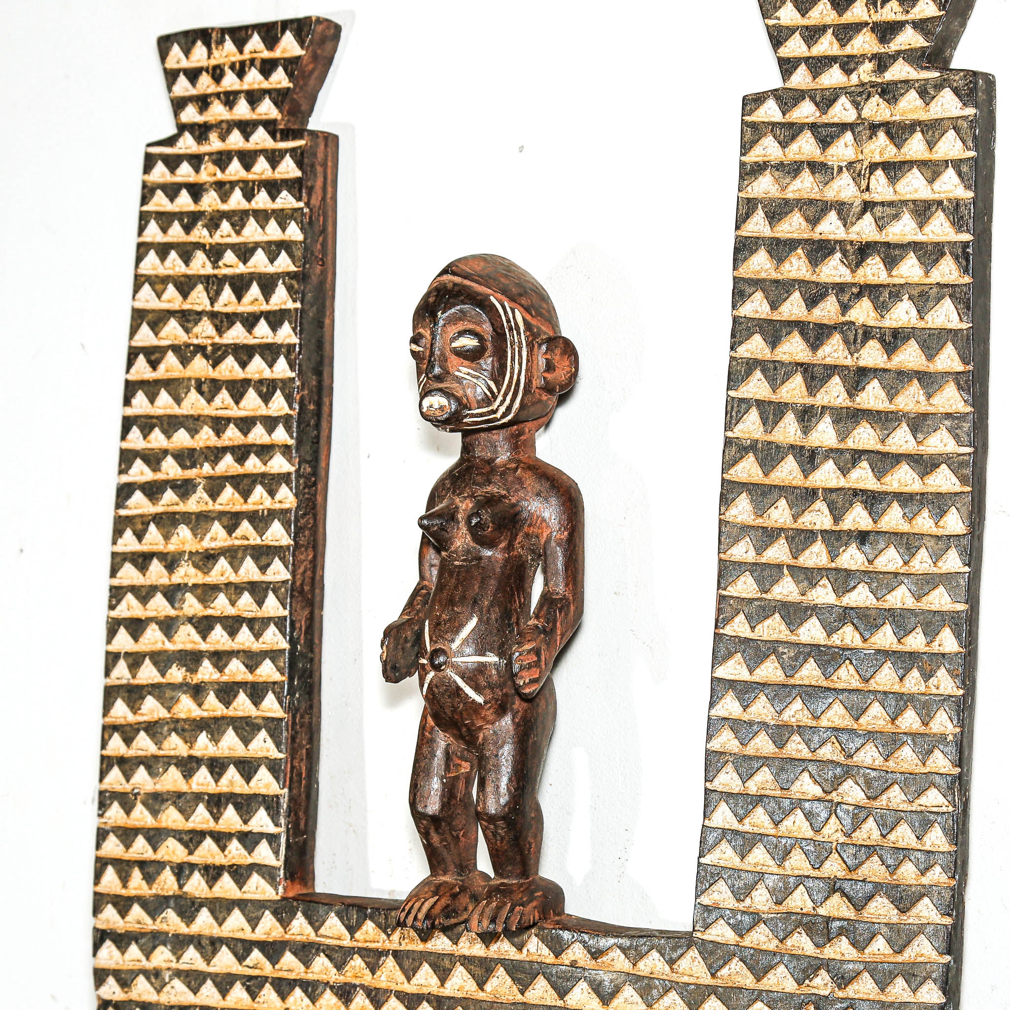 Wooden sculpture of a figure between two triangular patterns on a African mask on a white background