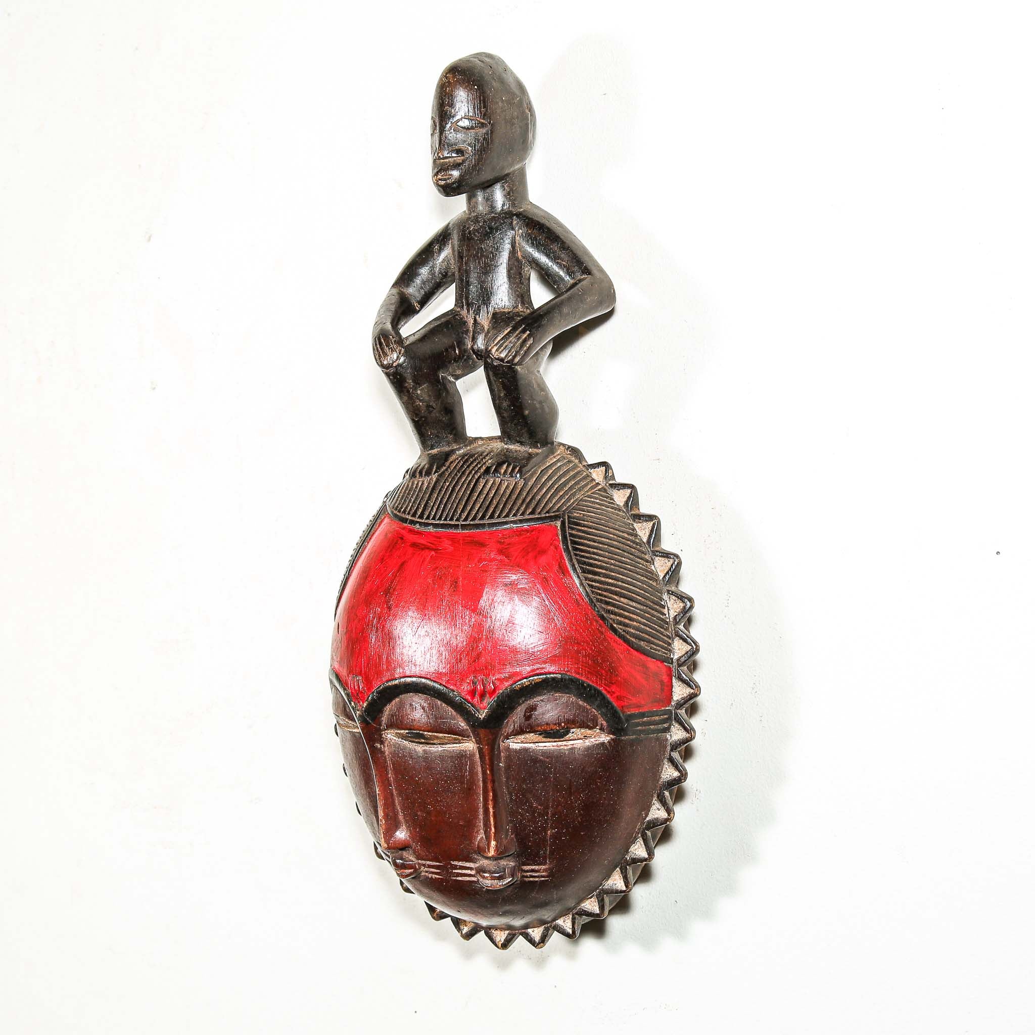 African mask with a red face and a figure on top against a white background