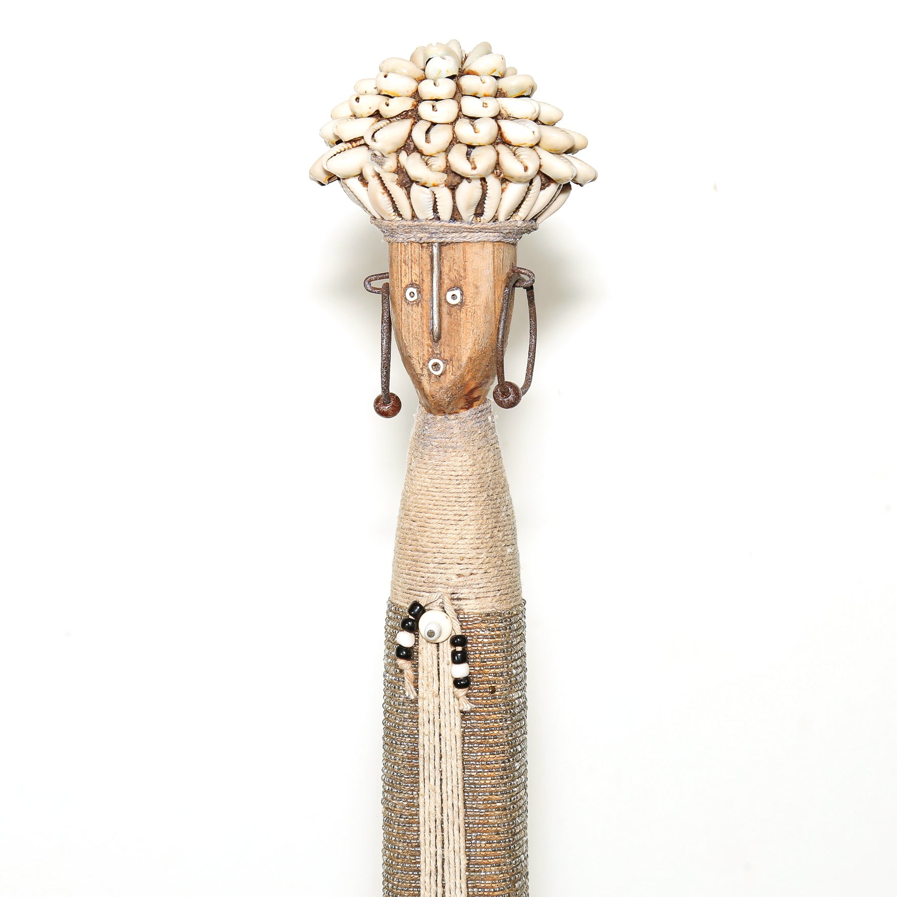 A handcrafted African-style wooden figurine of a woman with a shell-covered head, decorated with woven raffia, beads, and a long tassel, set against a plain white background.