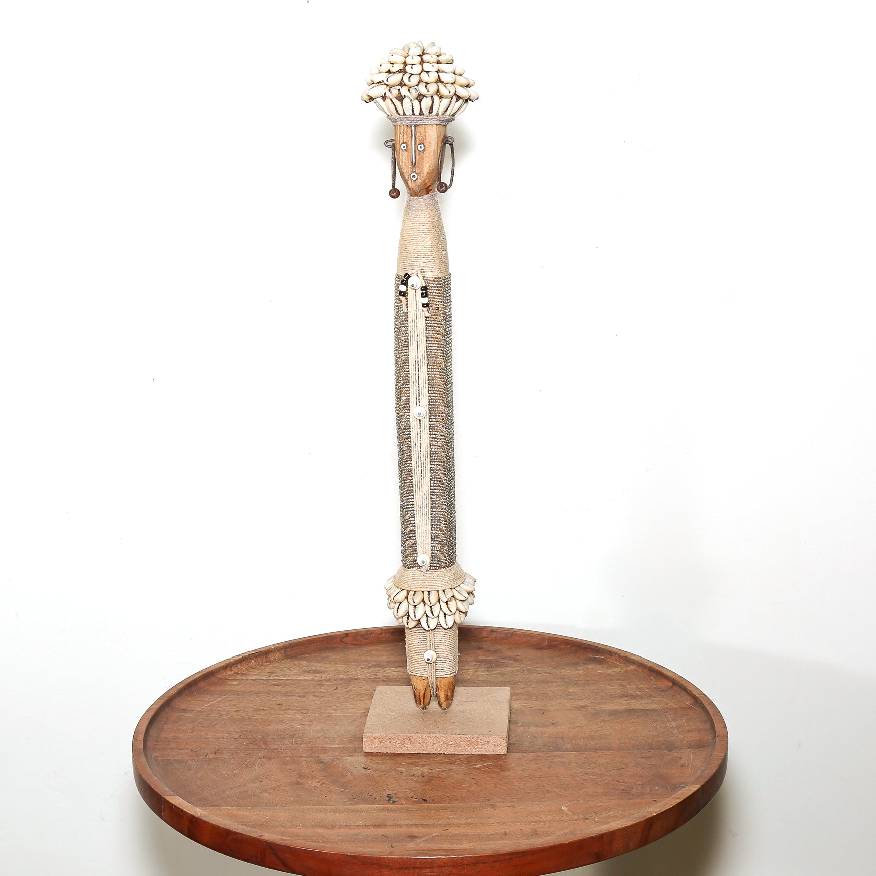 A handcrafted African-style wooden figurine with a tall, slender body, decorated with natural wood, shells, and beads, standing on a small square base atop a round wooden display platter against a white background.