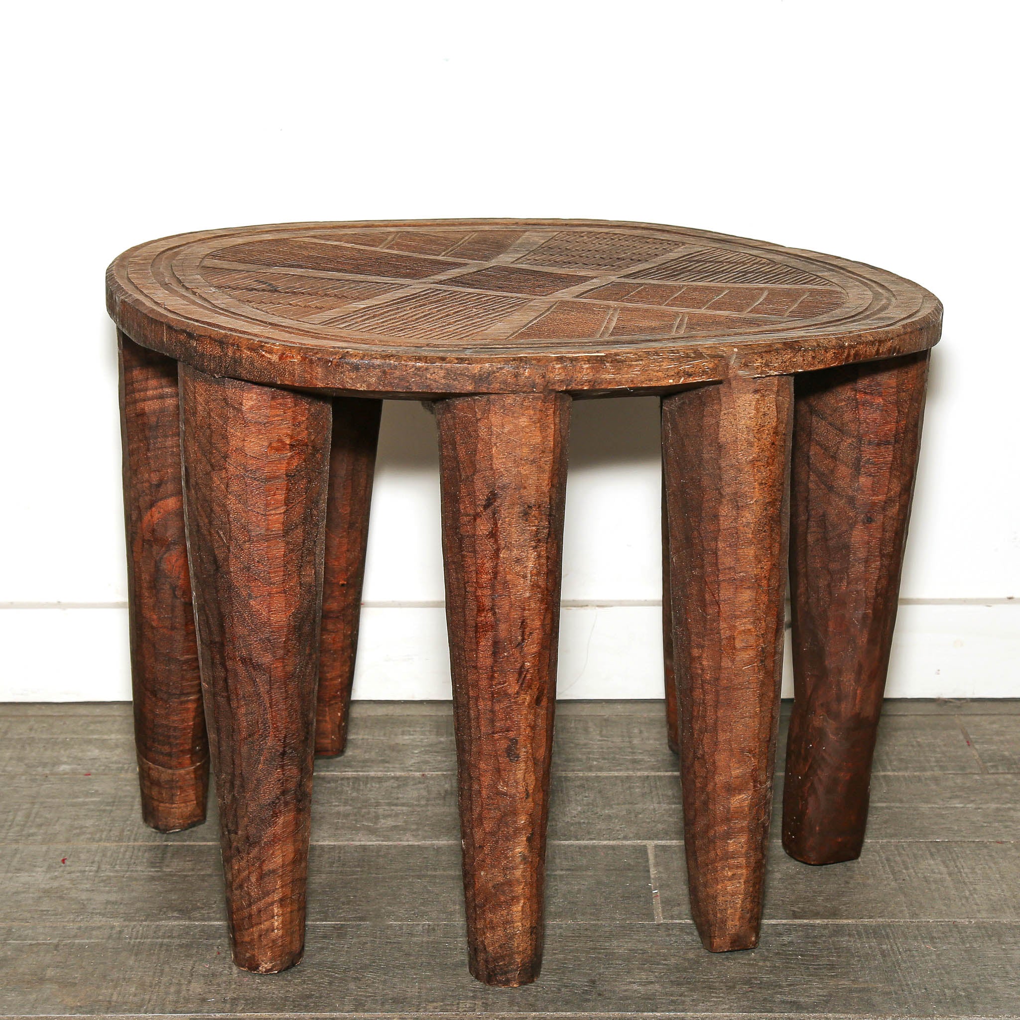 Round Nigerian Nupe stool with carved top and sculptural wooden legs. A rustic oval wooden stool with carved geometric patterns on the top and four thick, tapered legs, shown against a white wall and gray floor.