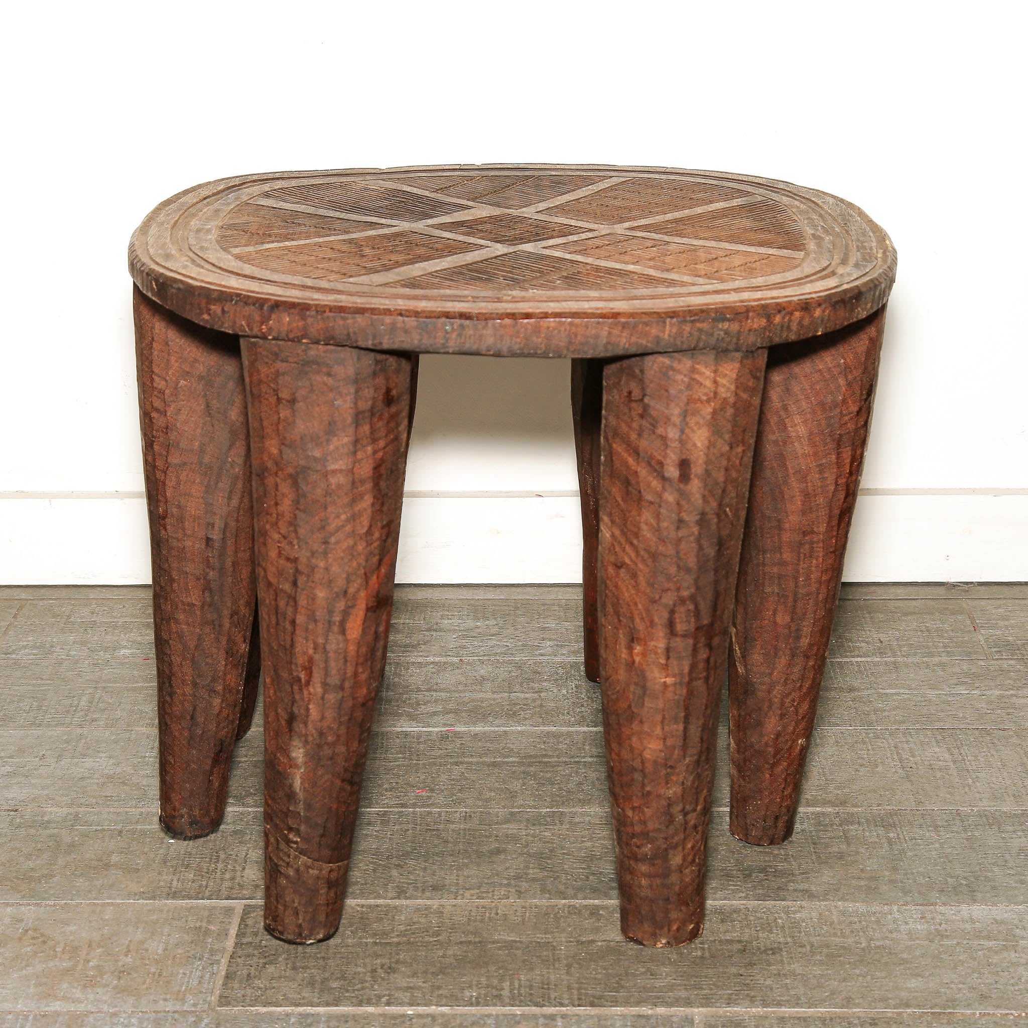 Hand-carved Nupe stool from Nigeria displayed as African decor-A rustic dark brown wooden stool with a round seat featuring a geometric inlay pattern and four sturdy, slightly tapered legs, set against a white wall and gray tiled floor.