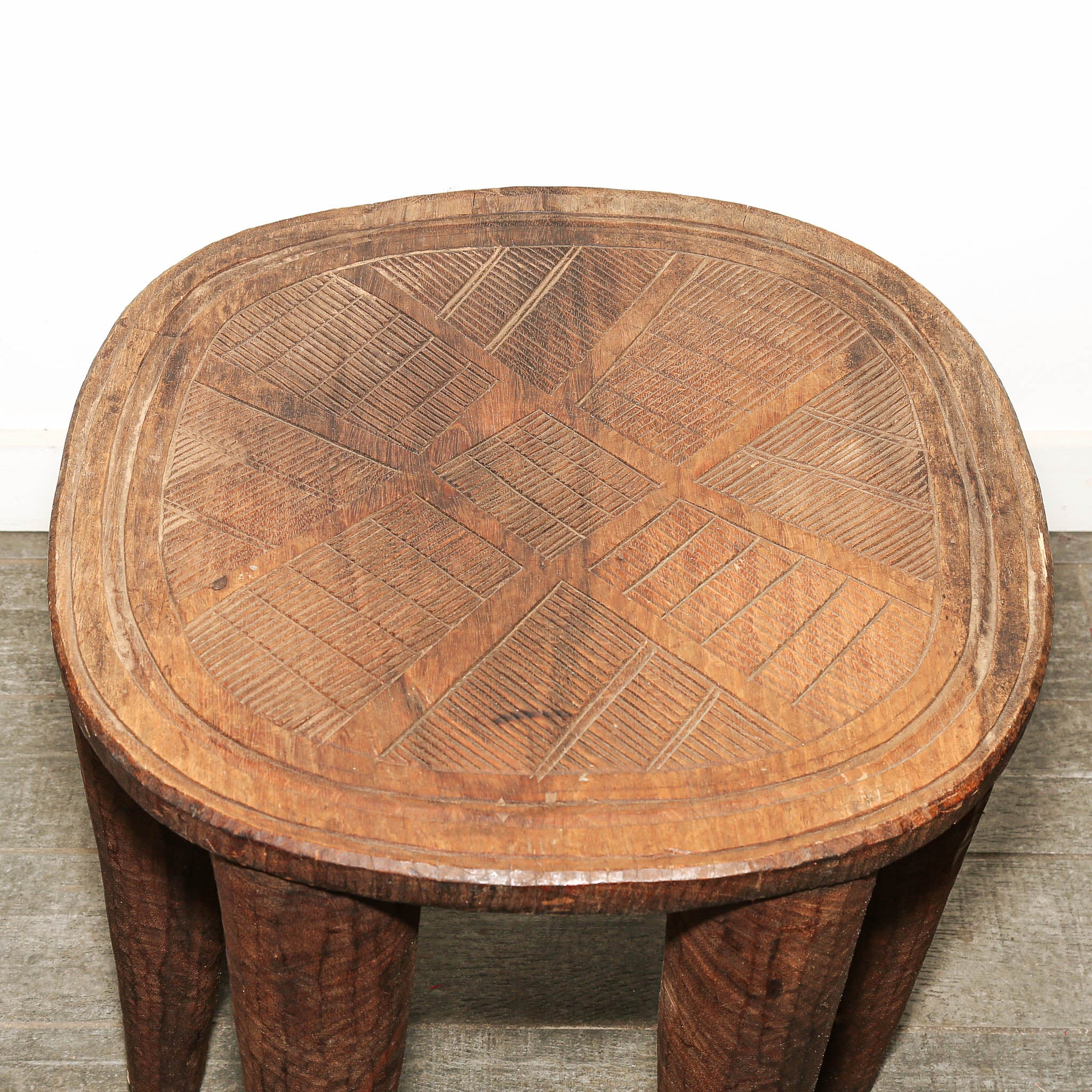 Traditional African Nupe wood stool carved from a single block of wood. A rustic carved wooden stool with a thick round seat and four legs, featuring geometric linear patterns on the top, set against a plain white wall and gray carpet.