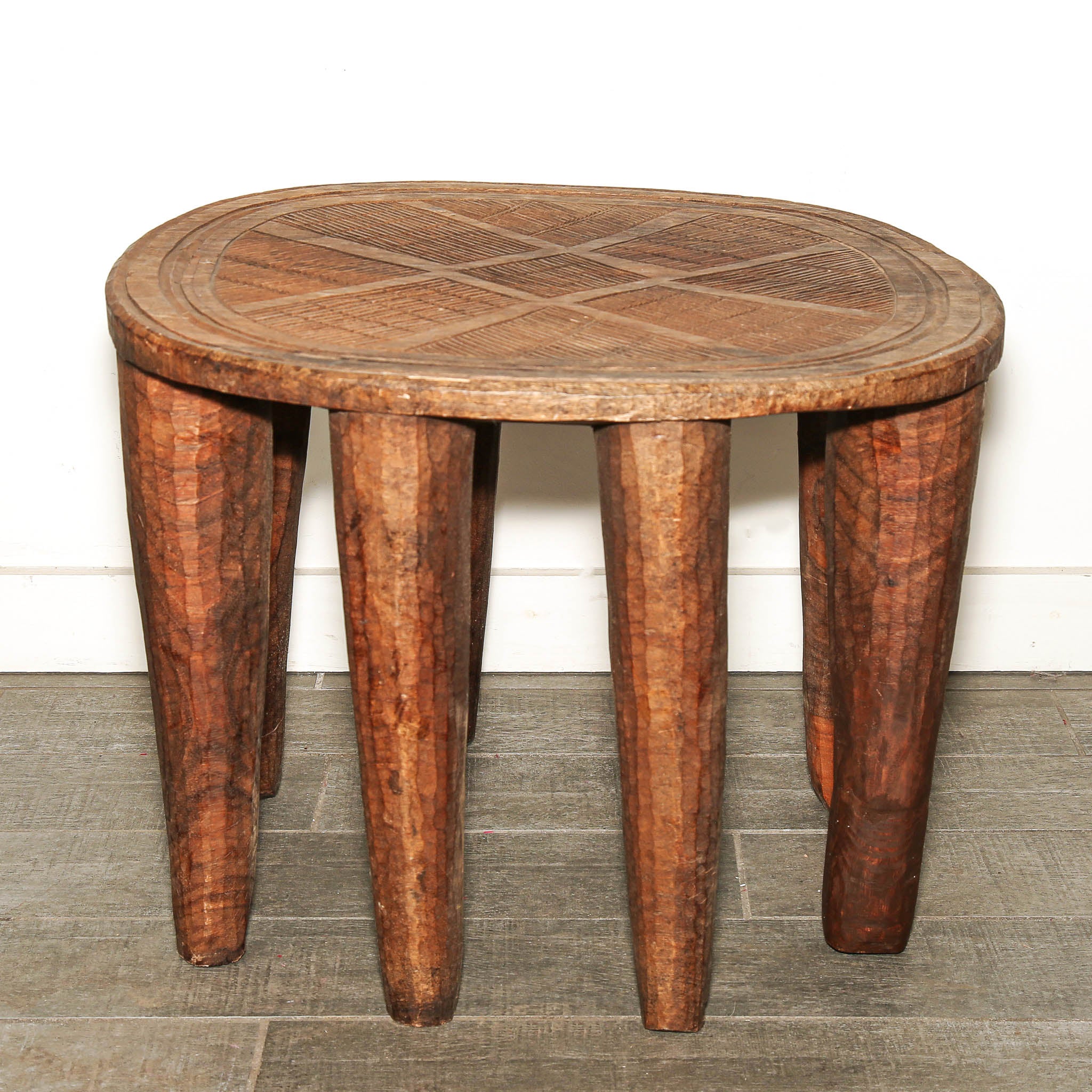 Large round Nupe stool from Nigeria with hand-carved geometric design. A rustic round wooden stool with many thick, textured legs and a geometric inlay pattern on the seat, set against a white wall and gray tiled floor.