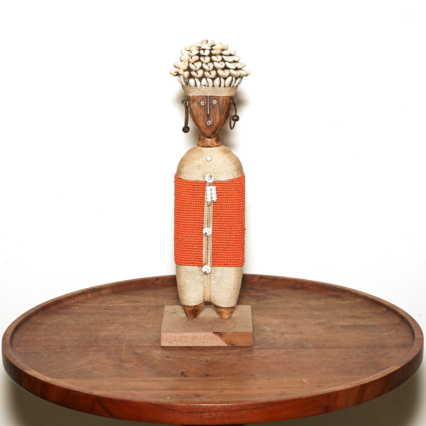 A handcrafted African wooden doll with a carved face, a woven orange and beige body, and a cluster of cowrie shells on its head, displayed on a round wooden tray against a white background.