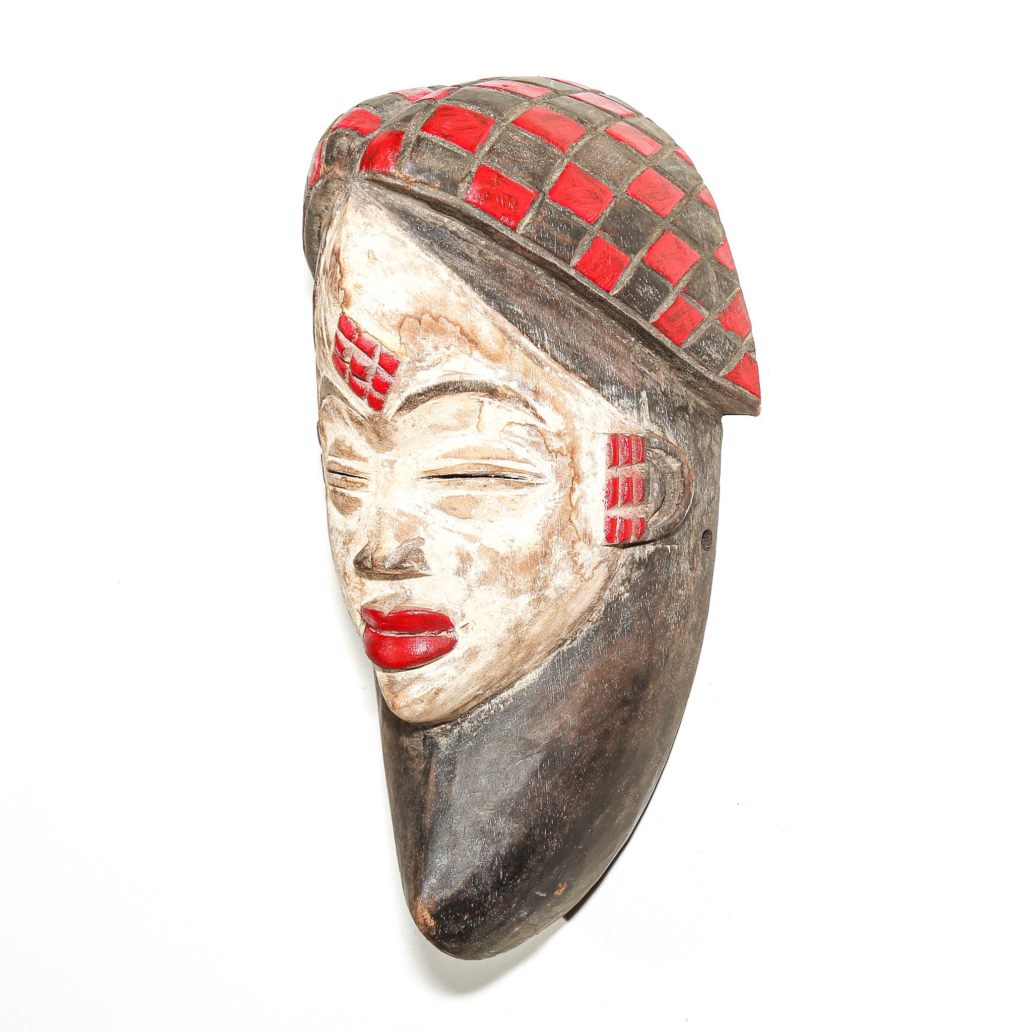 A carved African-style wooden mask depicting a woman's face in profile, featuring a red and dark checkered pattern on the hair, red painted details on the forehead and ear, and bright red lips, set against a plain white background.