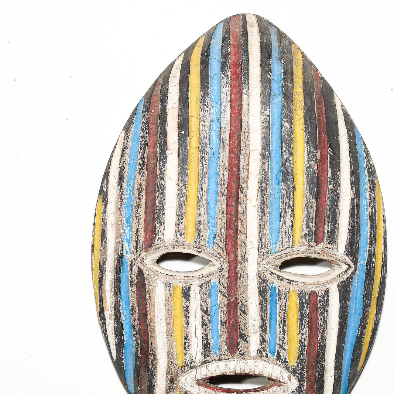 A close-up of a carved wooden mask with vertical painted stripes in blue, yellow, red, black, and off-white, featuring cut-out eyes and a mouth at the bottom.
