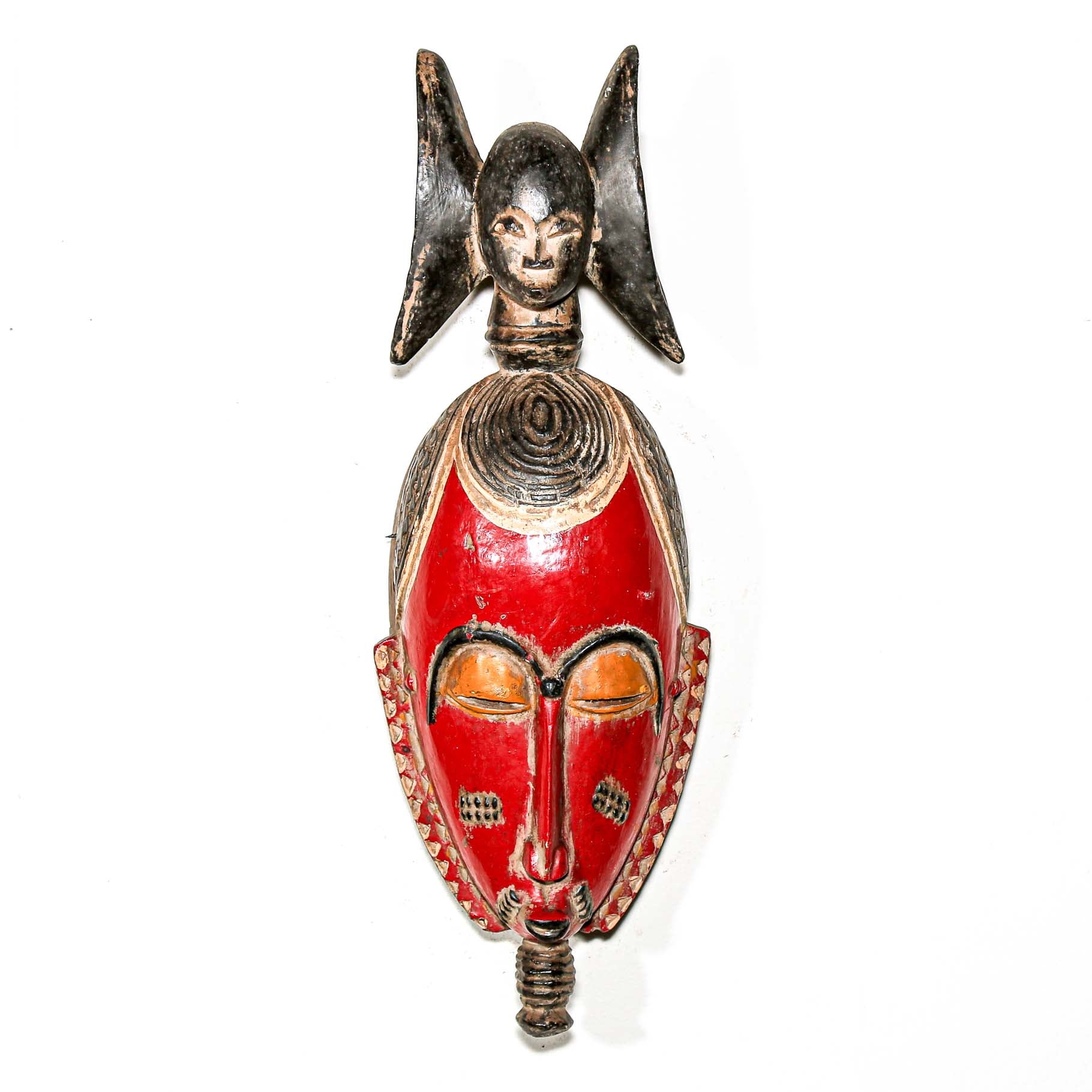 Hand-made African mask with red face and decorative black headdress on a white wall