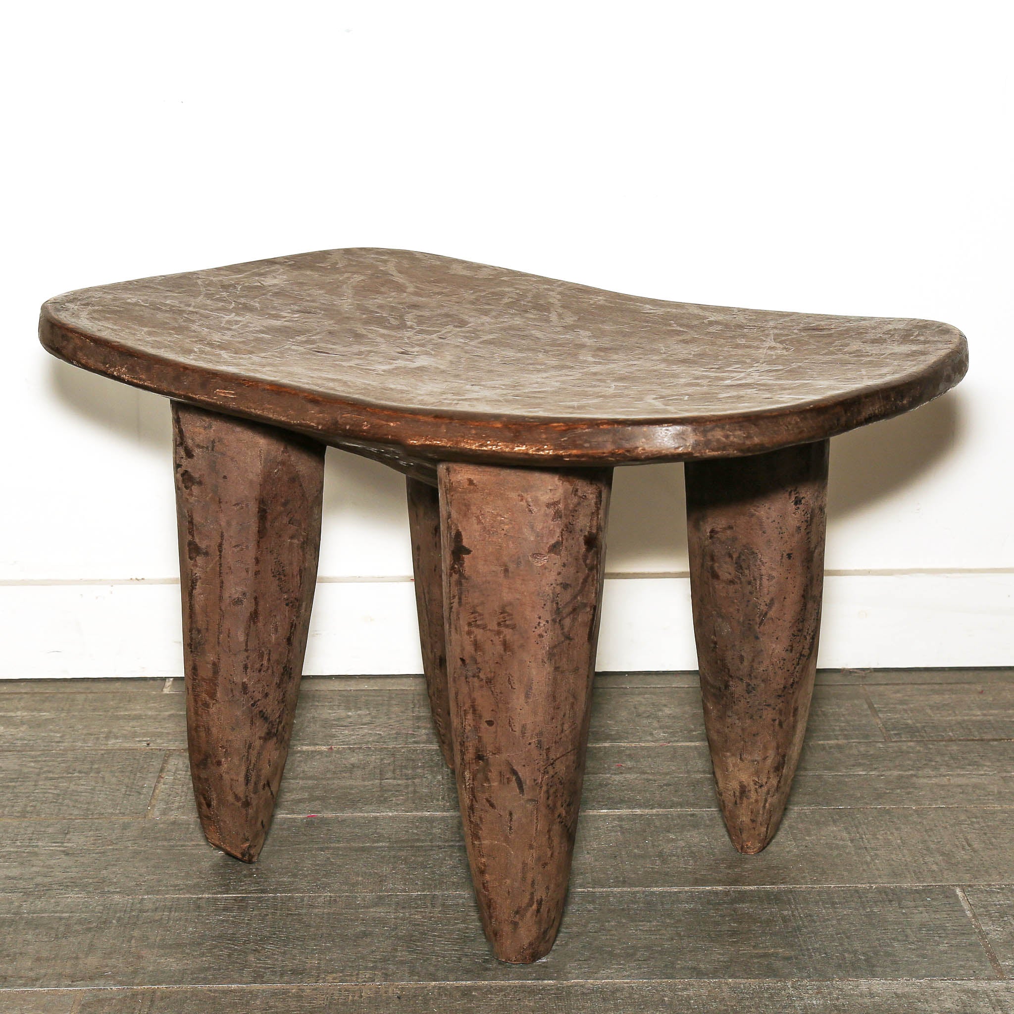 Senufo table from West Africa with curved wooden top and tapered legs and organic-shaped seat and four sturdy legs, shown on a gray wood floor against a white wall.