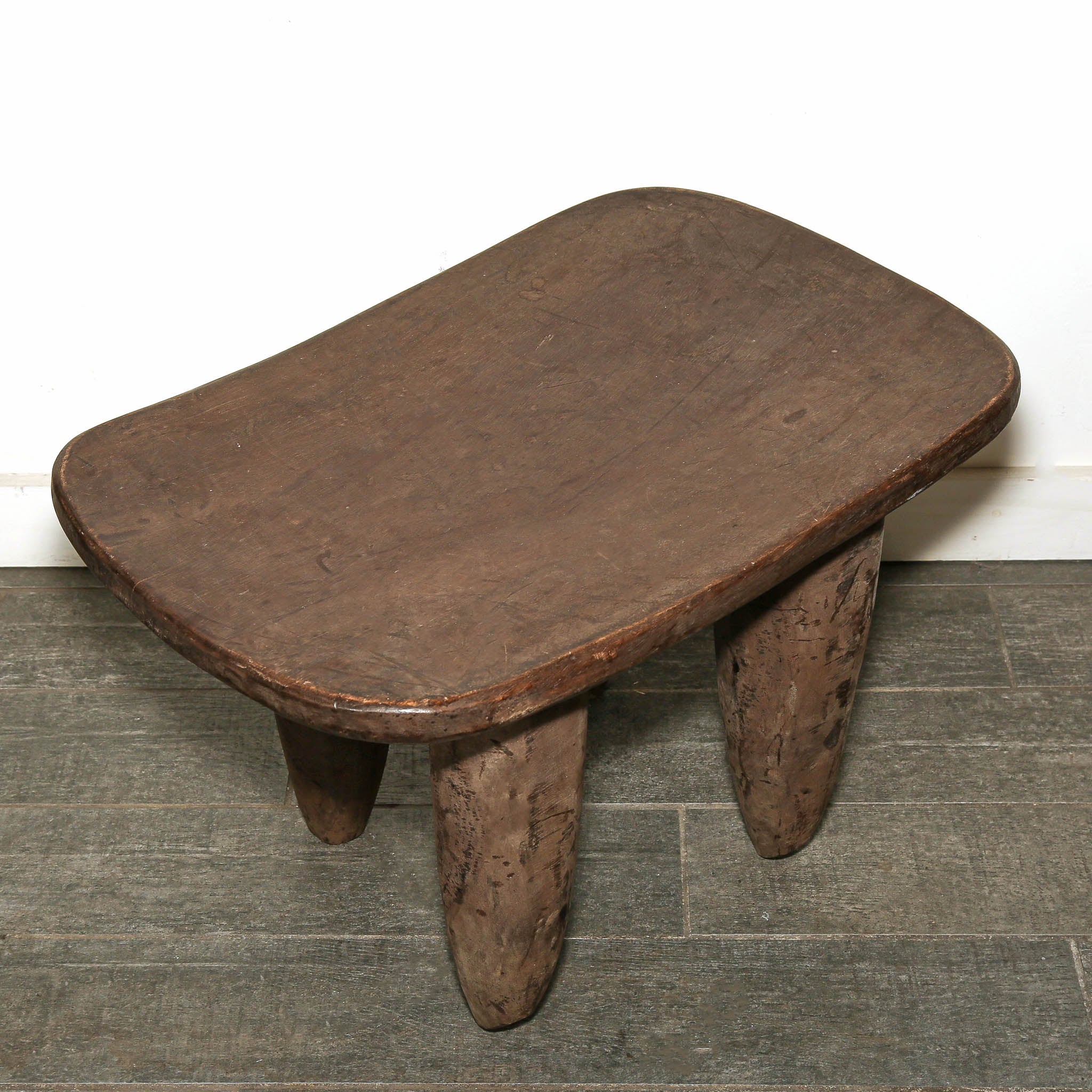Sculptural Senufo African table displayed as functional art and home decor-A rustic dark brown wooden stool with a thick rounded rectangular seat and three sturdy legs, set against a white wall and gray tiled floor.