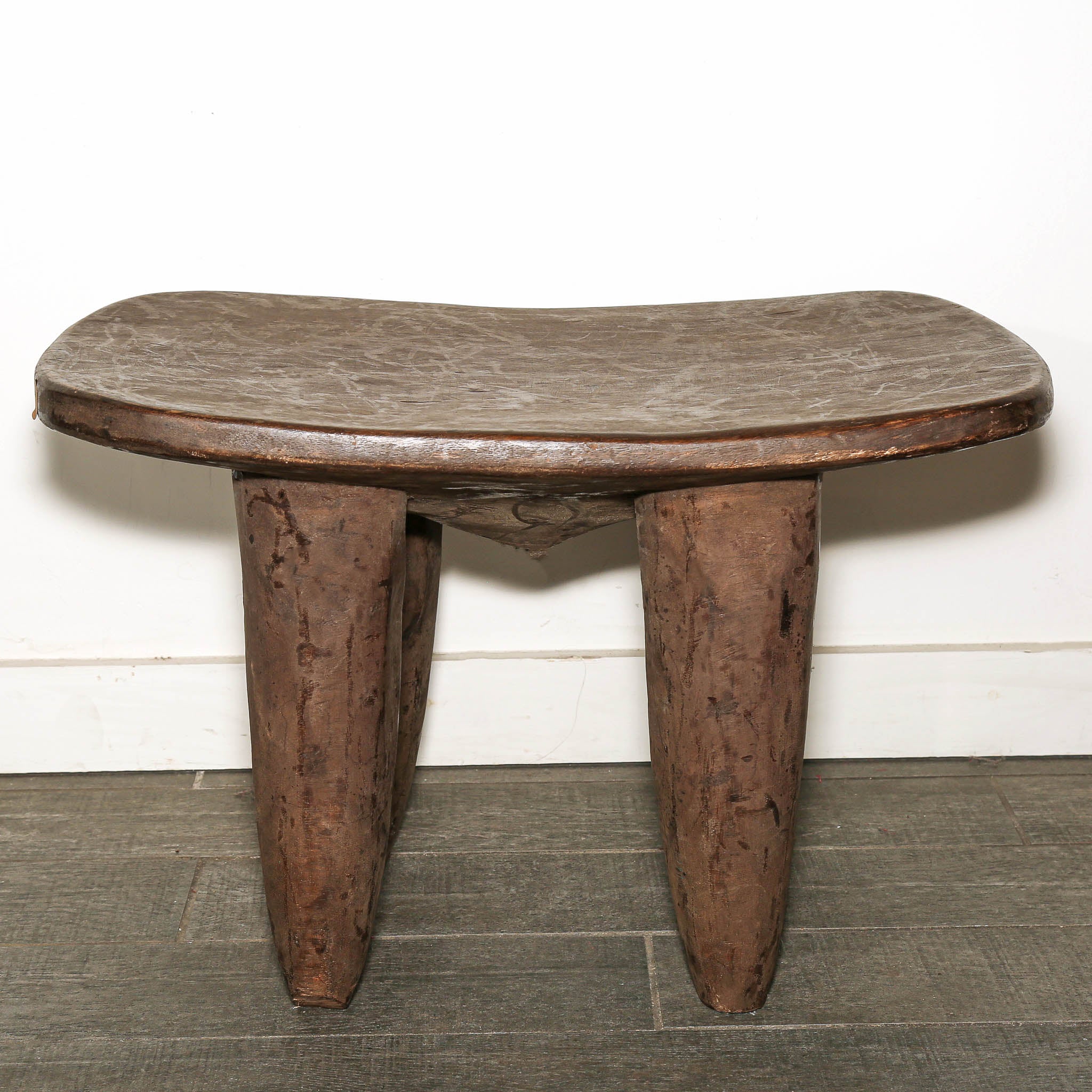 Hand-carved Senufo wood table inspired by traditional textile washboards. A rustic carved wooden stool with a curved, thick seat and three sturdy legs, set against a white wall and gray tiled floor.