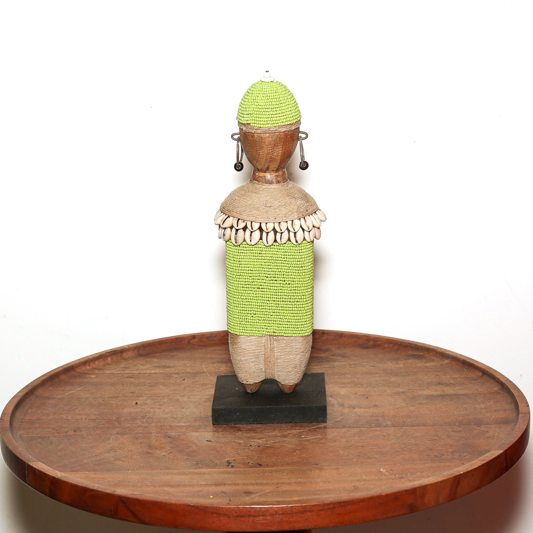 A handmade African-style wooden doll figurine with a bright green beaded wrap, a natural wood body, and cowrie shells around the neck, displayed on a round wooden table against a white background.