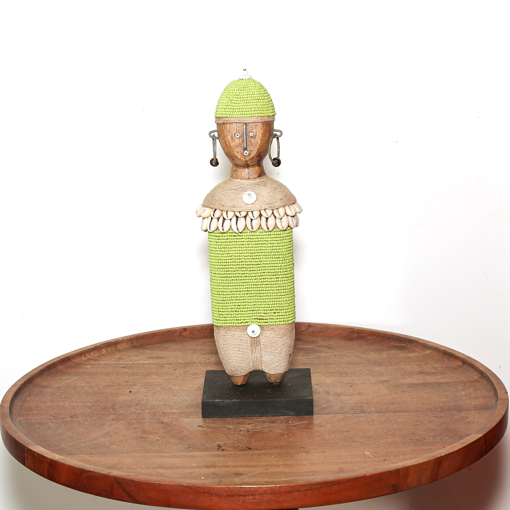 A handmade African-style wooden figurine with a green beaded body and hat, mounted on a black base and displayed on a round wooden tray against a white background.