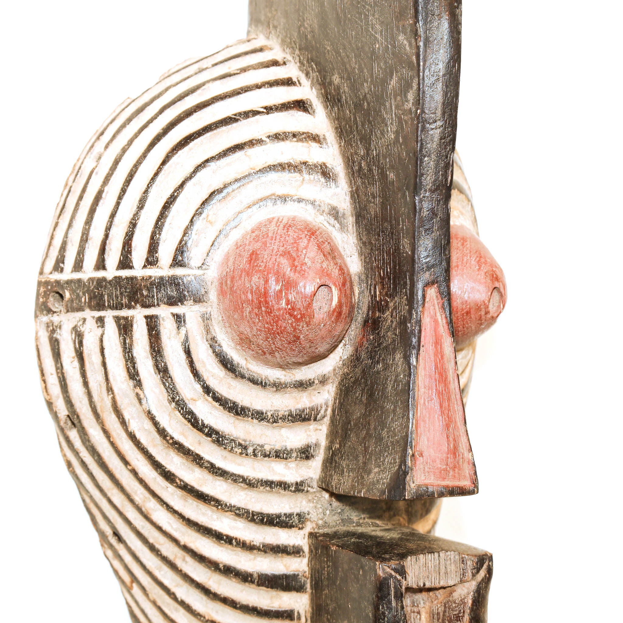 A close-up of a carved wooden mask with black and white concentric line patterns, reddish-brown painted eyes, and a reddish-brown nose on a white background.