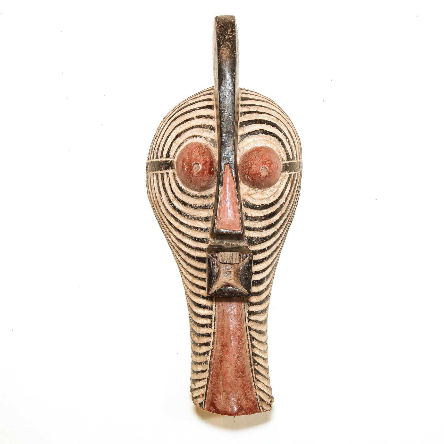 A carved wooden African-style mask with a long vertical form, featuring black concentric line patterns, reddish-brown painted areas on the eyes and lower face, and a dark top loop, shown against a plain white background.