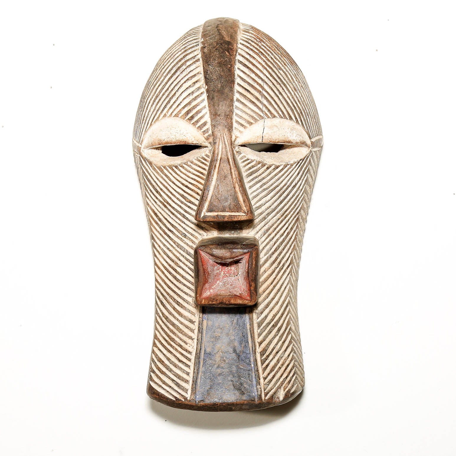 An African-style carved wooden mask with a textured, ribbed surface, featuring a reddish-brown square mouth and a blue-gray vertical accent near the bottom, set against a plain white background.