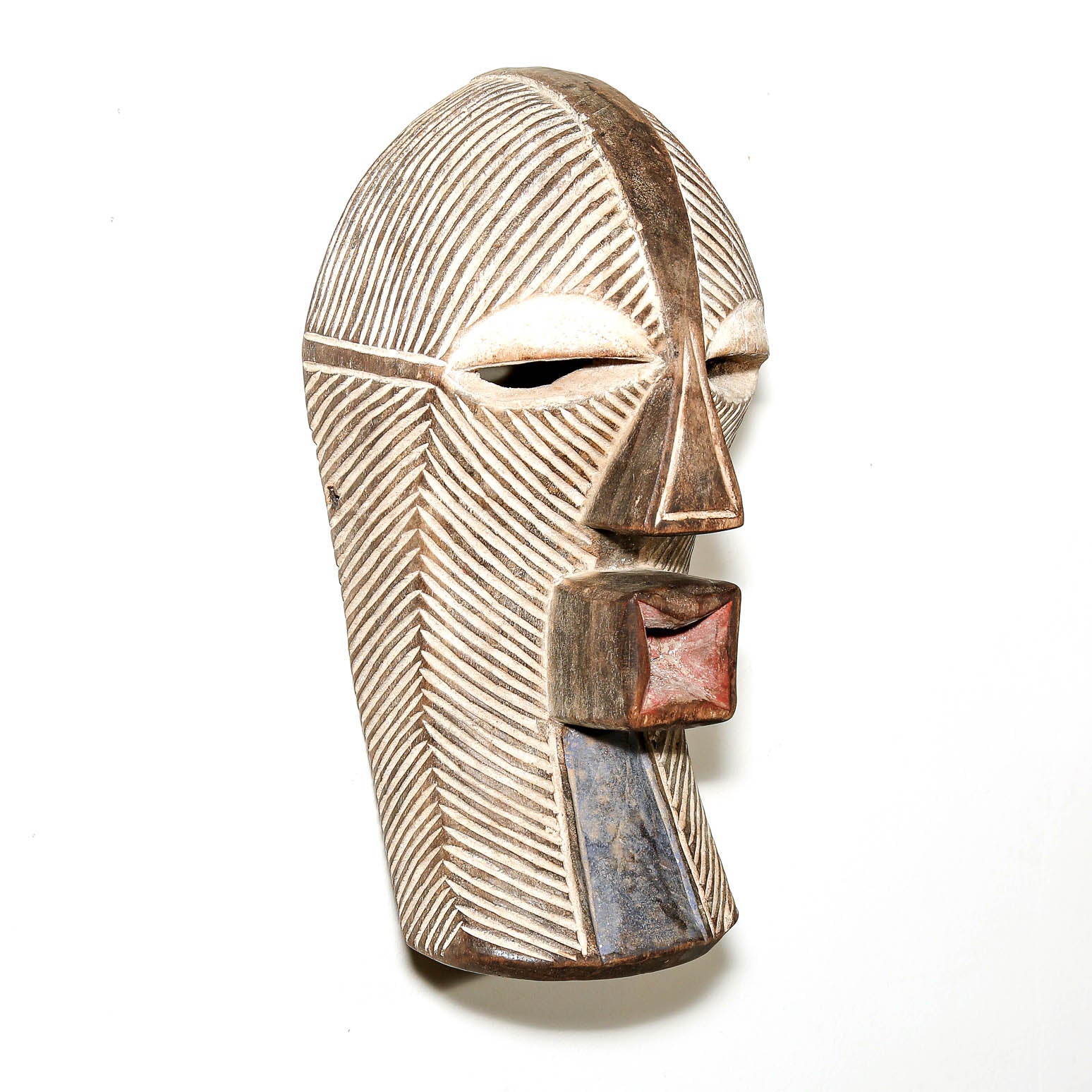 A carved wooden mask with geometric facial features, textured diagonal grooves, and earthy brown, beige, and blue painted accents, photographed against a plain white background.