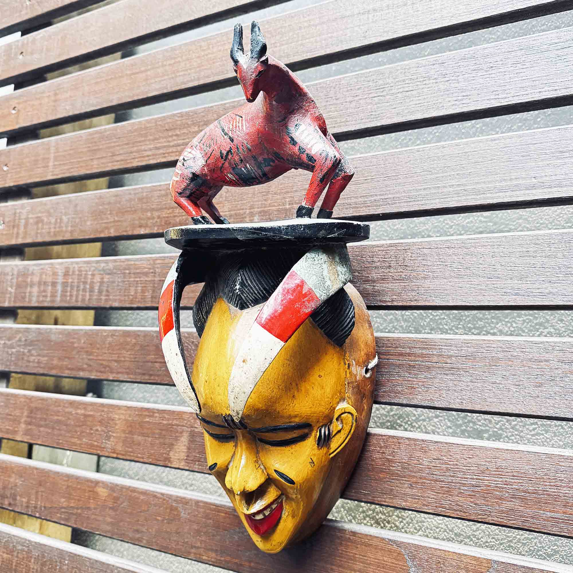 mask from africa with a yellow face and with a red full figure on top, against a wooden slat background