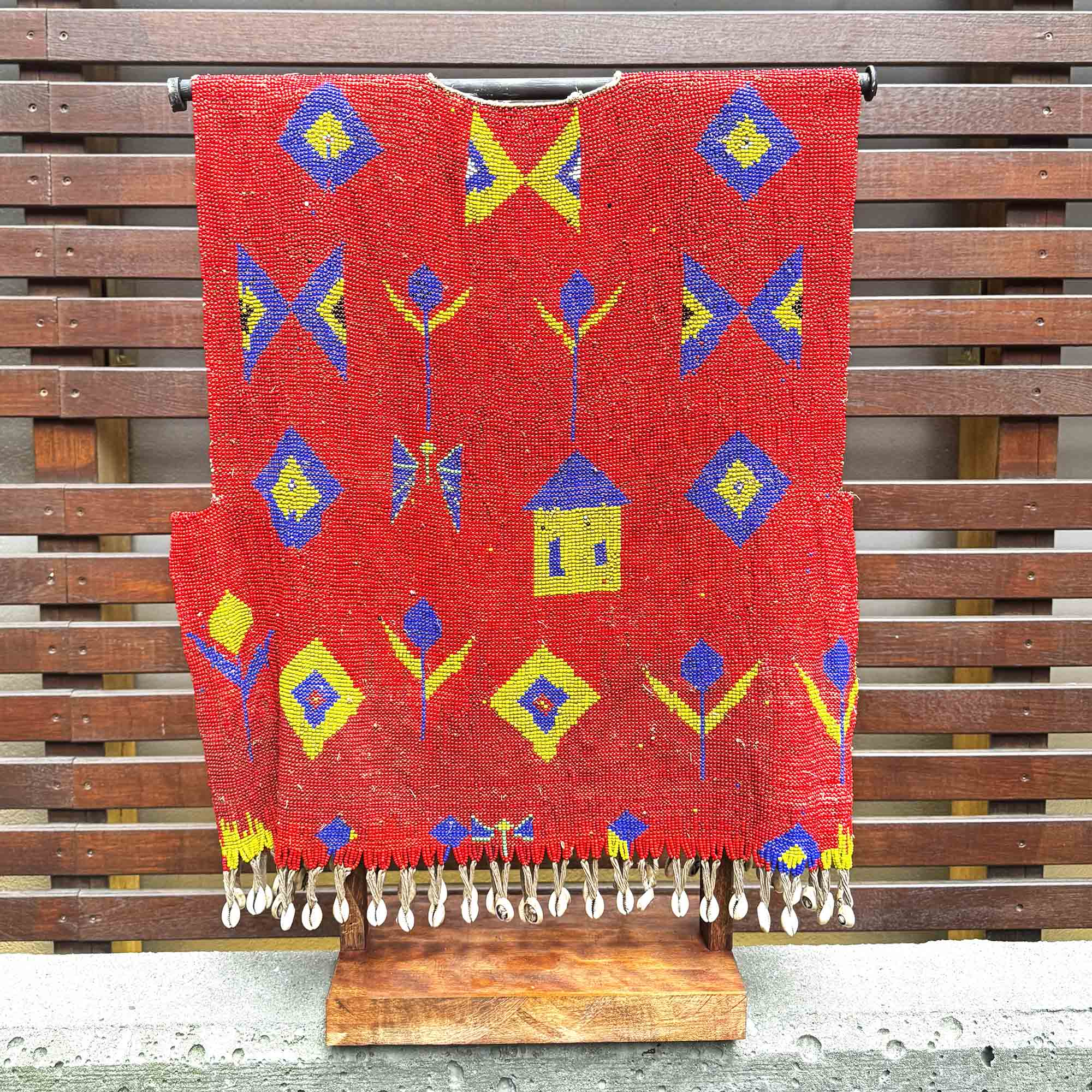 Red woven with beads african shirt with colorful patterns hanging on a wooden stand against a wooden background