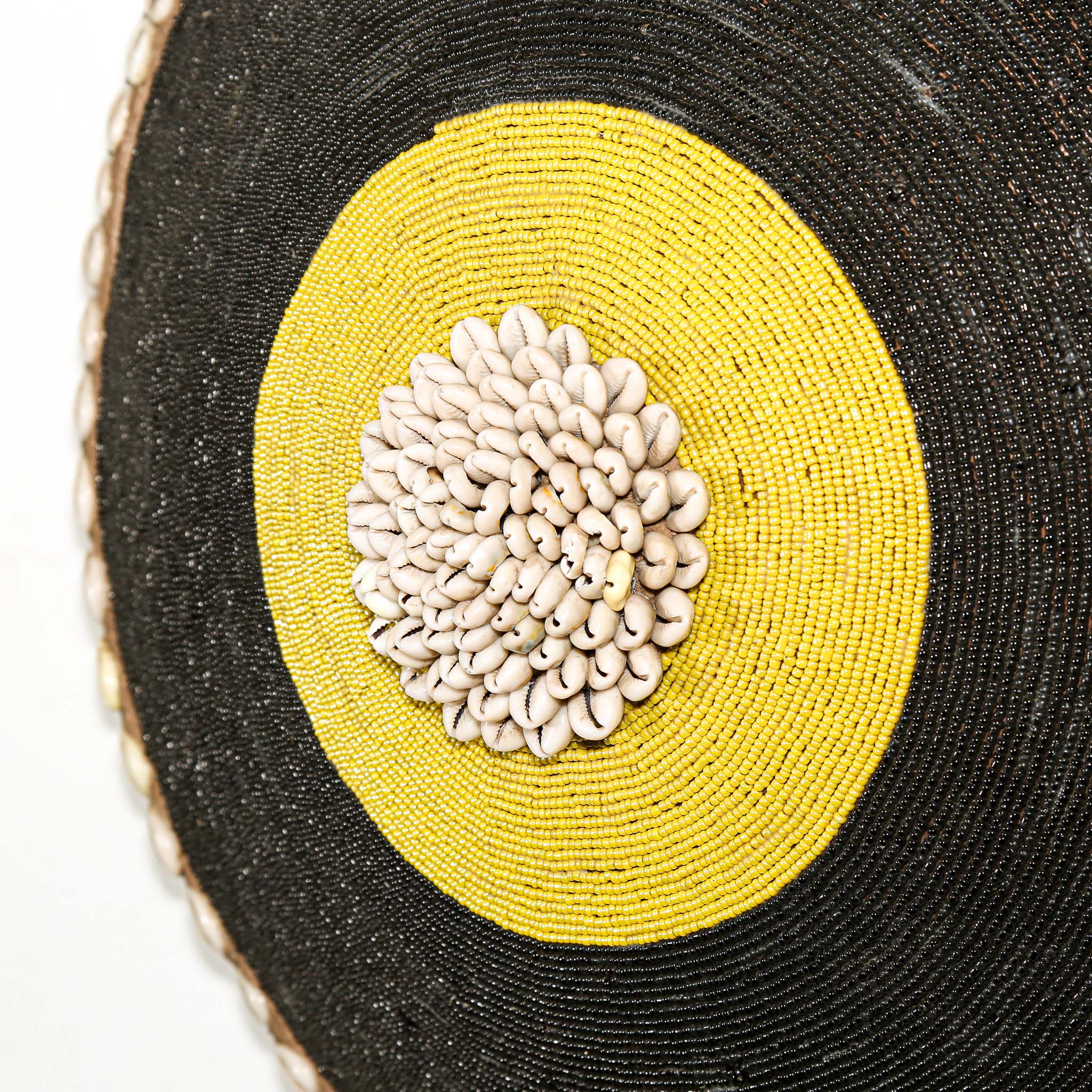 Round woven beaded shield with black surface, yellow center, and decorative edge hanging on a white wall