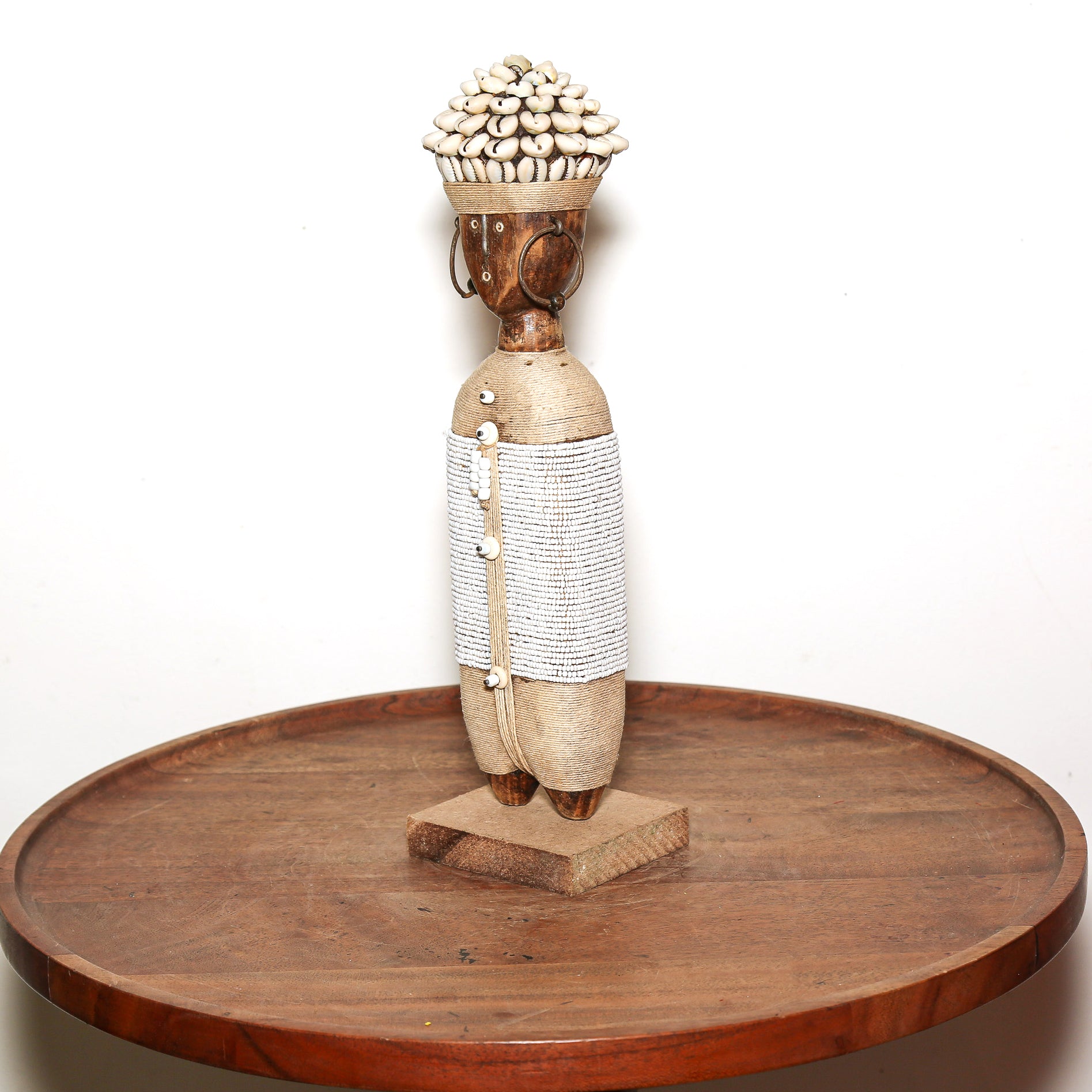 A handcrafted African-style wooden figurine with a woven body and a shell-covered head, displayed on a round wooden table against a plain white background.