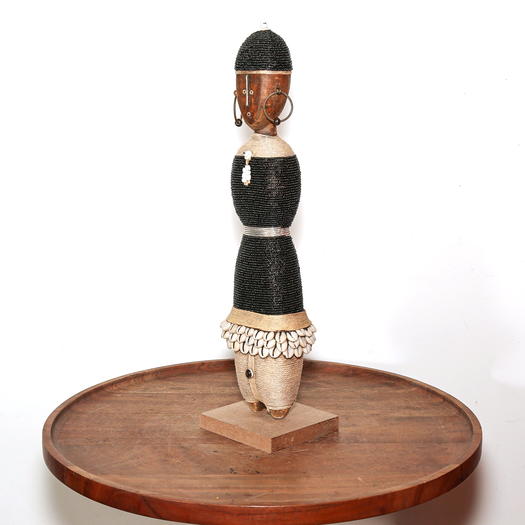 A traditional African wooden doll sculpture with a black woven dress, shell skirt, and wooden head, displayed on a round wooden platform against a white background.
