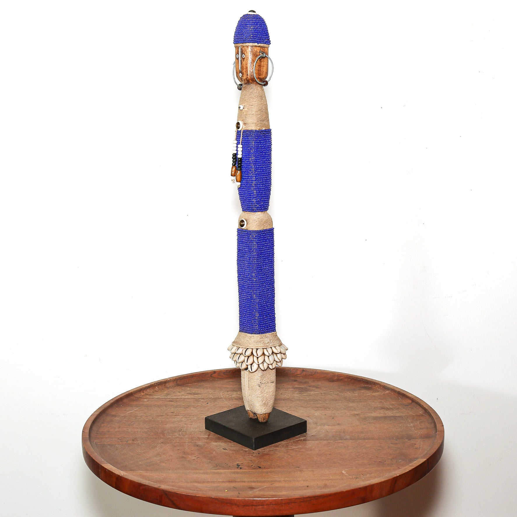 A tall, hand-carved wooden African-style doll figurine with blue wrapped sections, displayed on a round wooden stand against a white background.