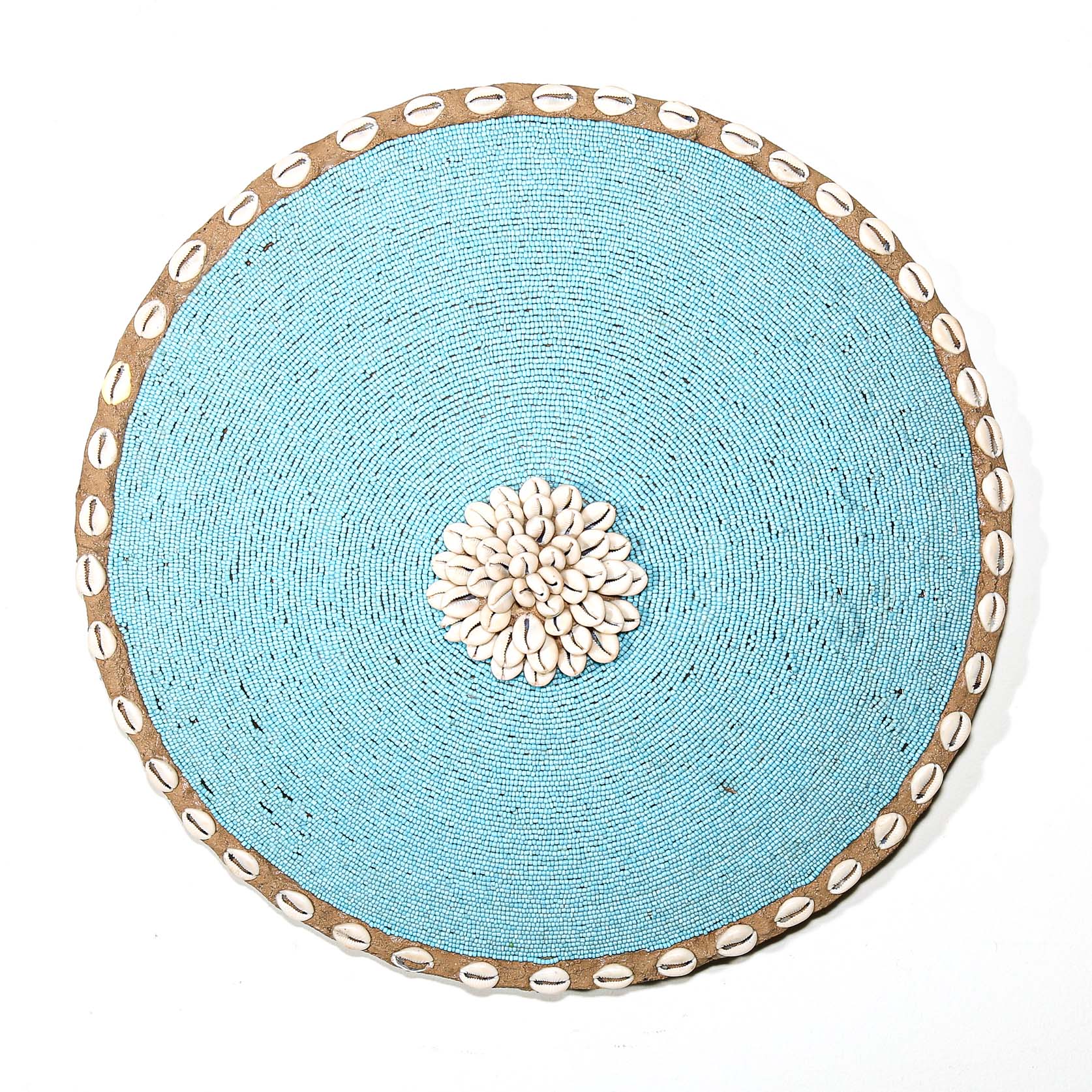 Round blue beaded wall shield decor with a central white flower design made from cowrie shells hanging on a white wall.