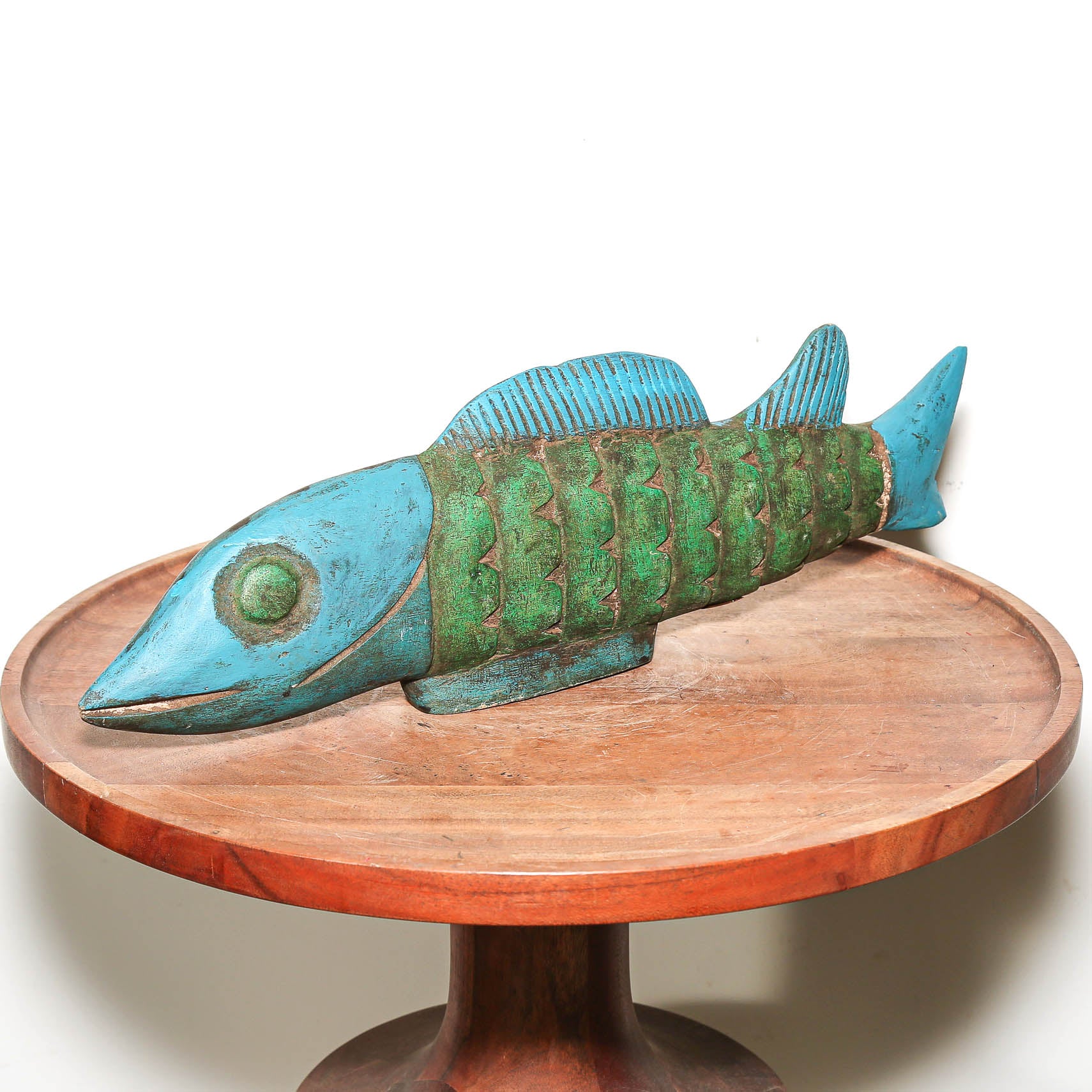 A decorative fish sculpture with a turquoise blue head and fin, and a green scaled body, displayed on a round wooden pedestal against a plain white background.