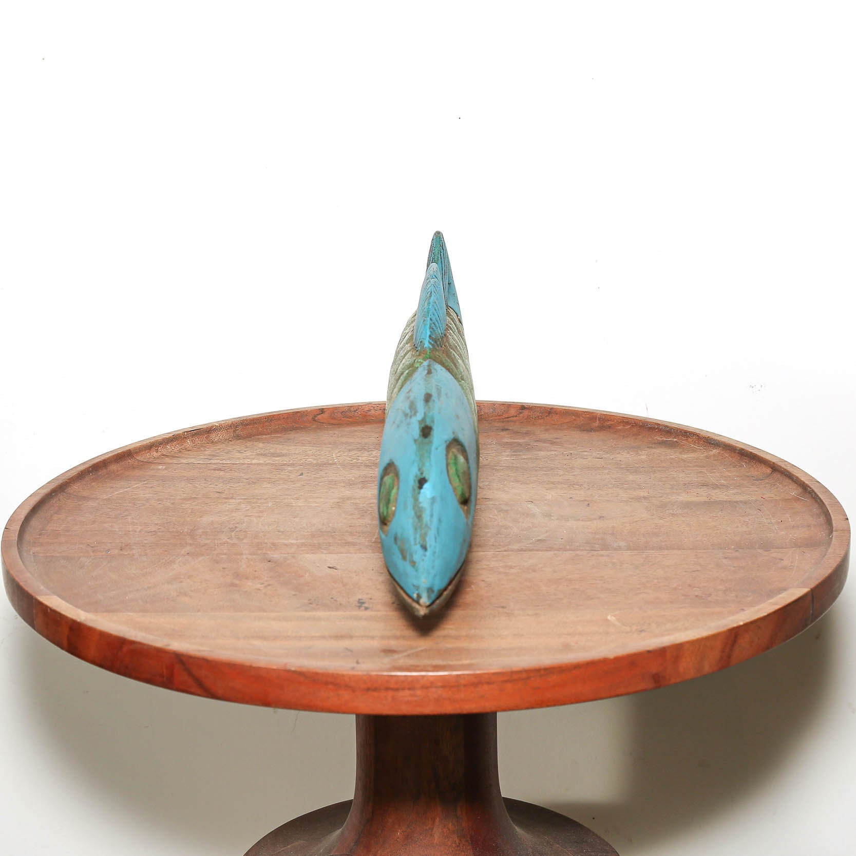 A small turquoise and green decorative object standing upright on a round wooden pedestal table against a plain white background.