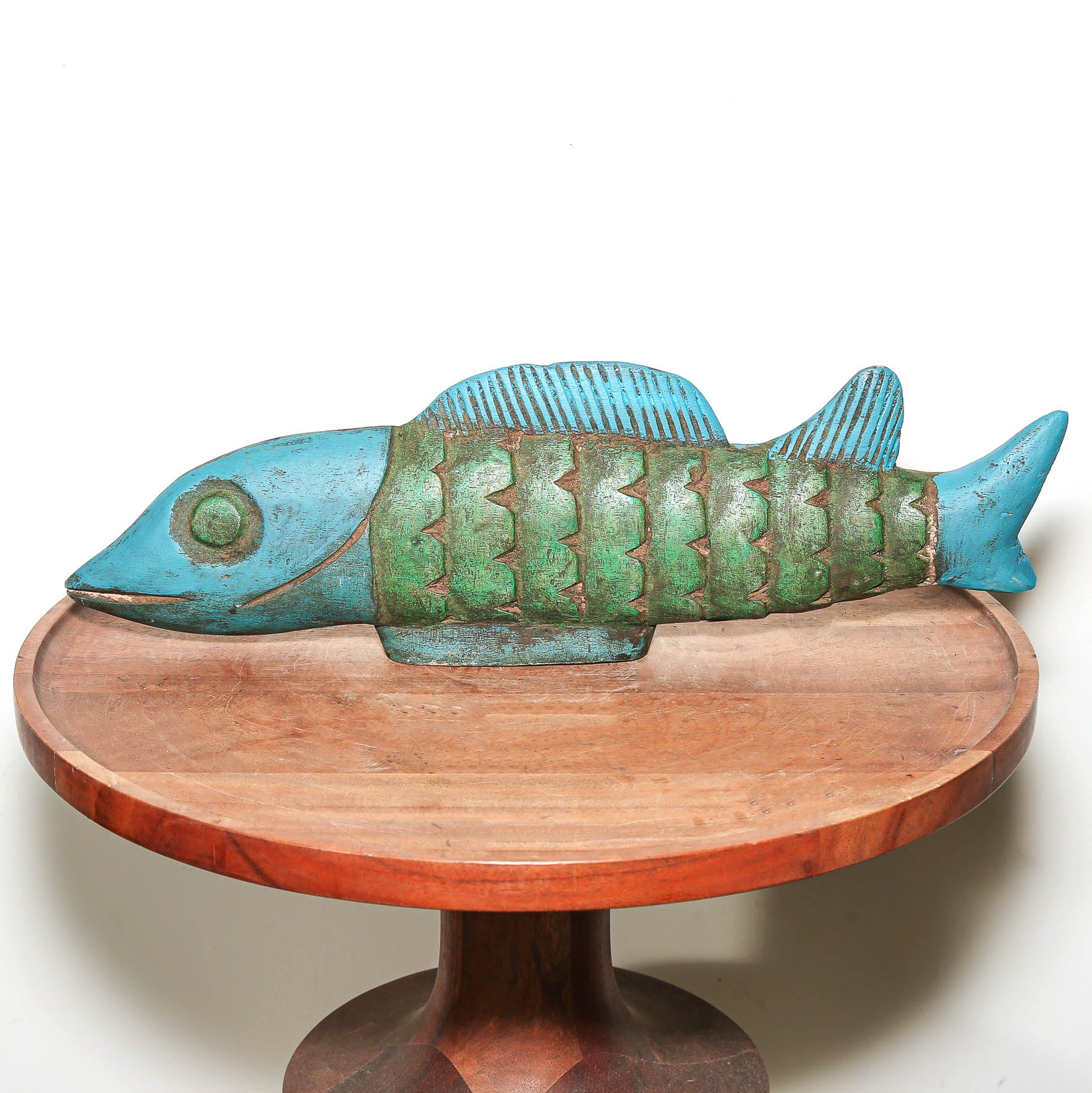 A decorative fish sculpture with a turquoise-blue head, eye, and fins, and a green body with textured scale patterns, displayed on a round wooden pedestal against a white background.