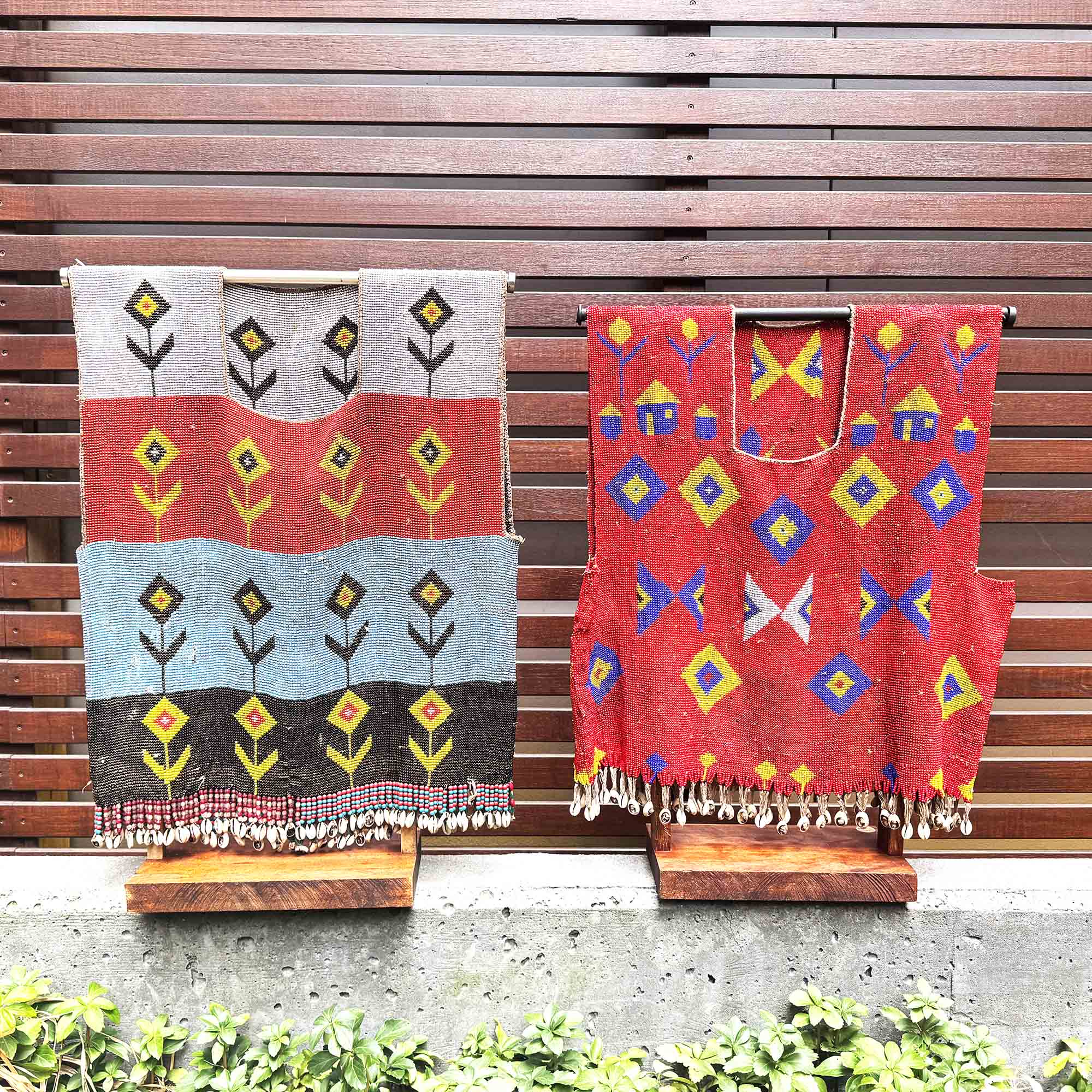 Two colorful african shirts sculpture with geometric patterns on stands against a wooden slat background.