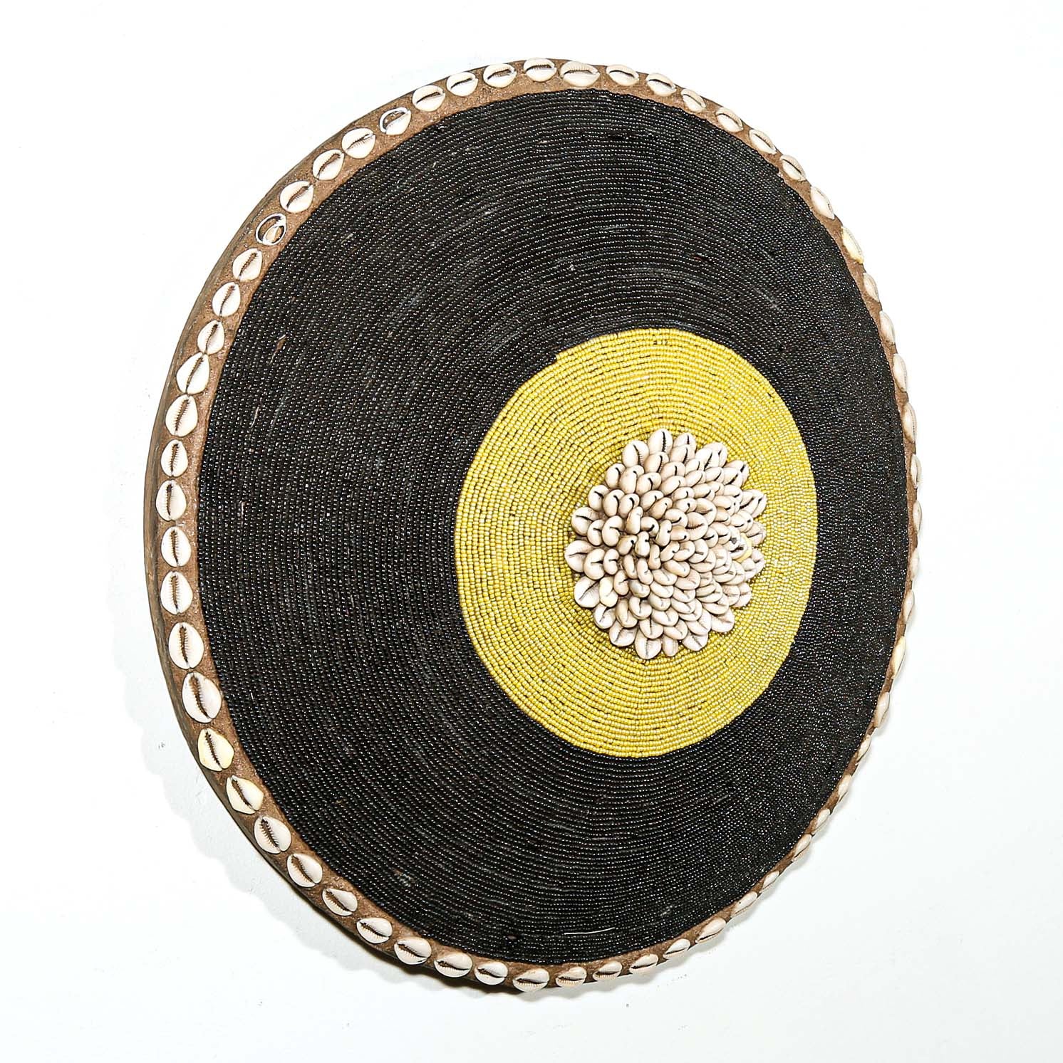 Round woven beaded shield with black surface, yellow center, and decorative edge hanging on a white wall