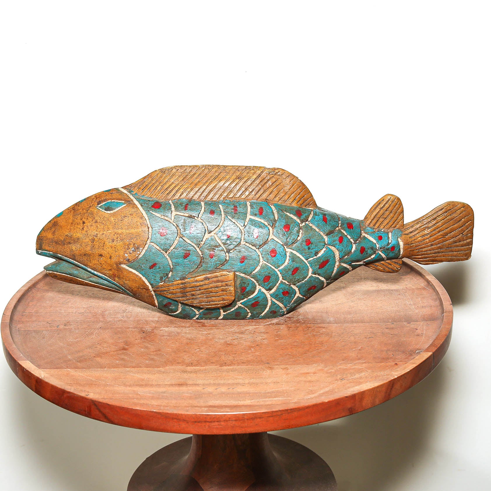 A hand-carved wooden fish sculpture with a teal, red, and gold painted scale pattern, displayed on a round wooden pedestal against a white background.