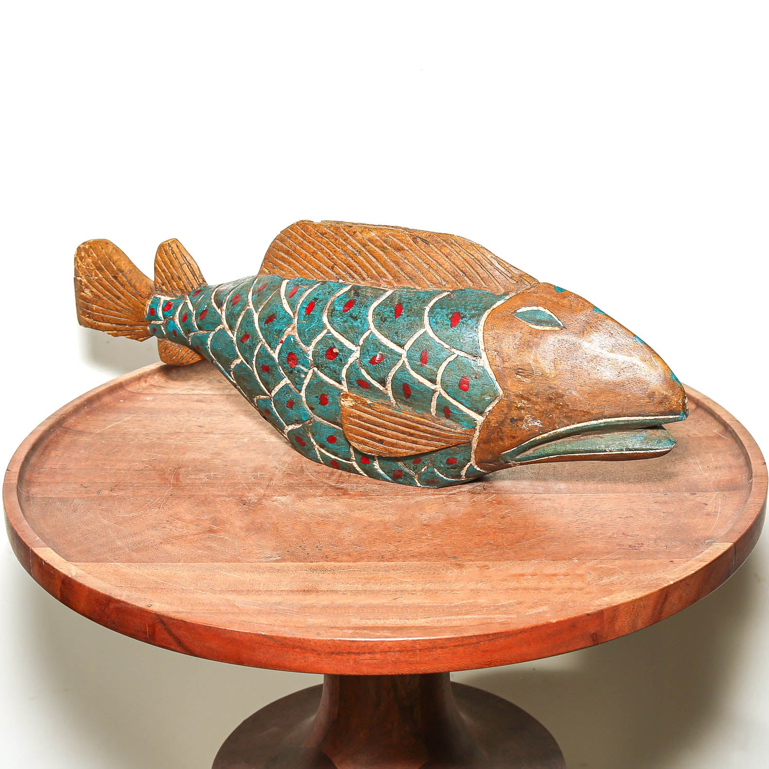 A hand-carved wooden fish sculpture with a teal and brown patterned body, displayed on a round wooden pedestal table against a white background.