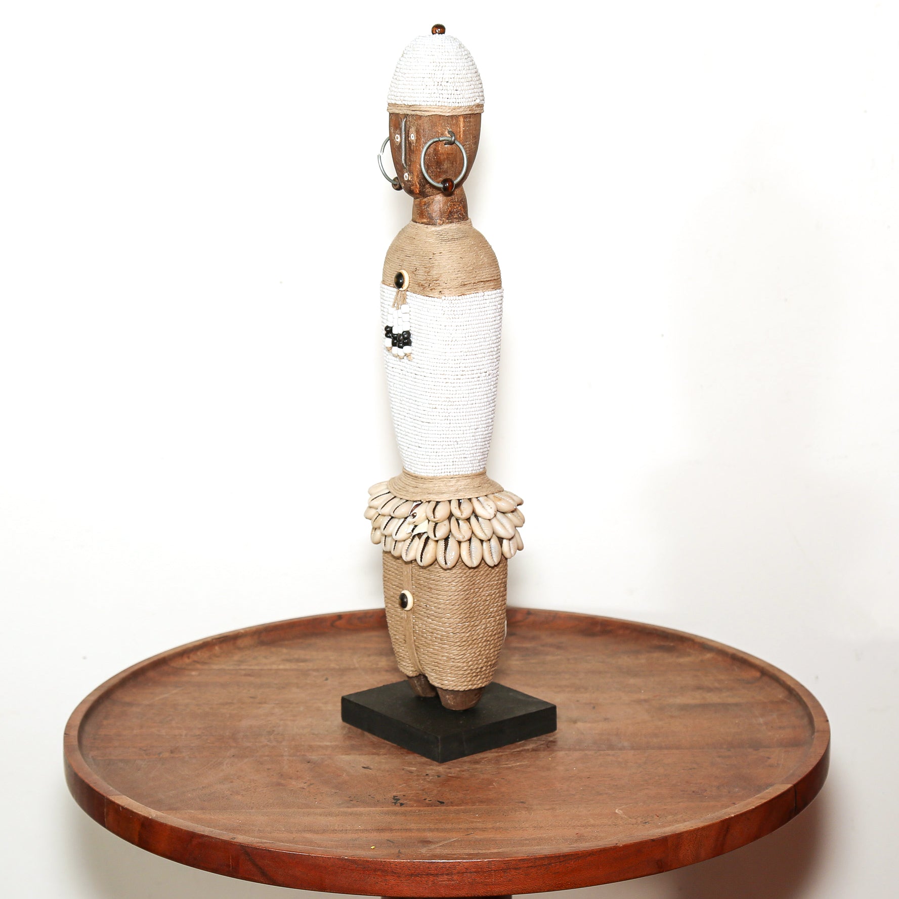A tall African-style wooden sculpture with woven white and natural wood sections, displayed on a round wooden table against a plain white background.