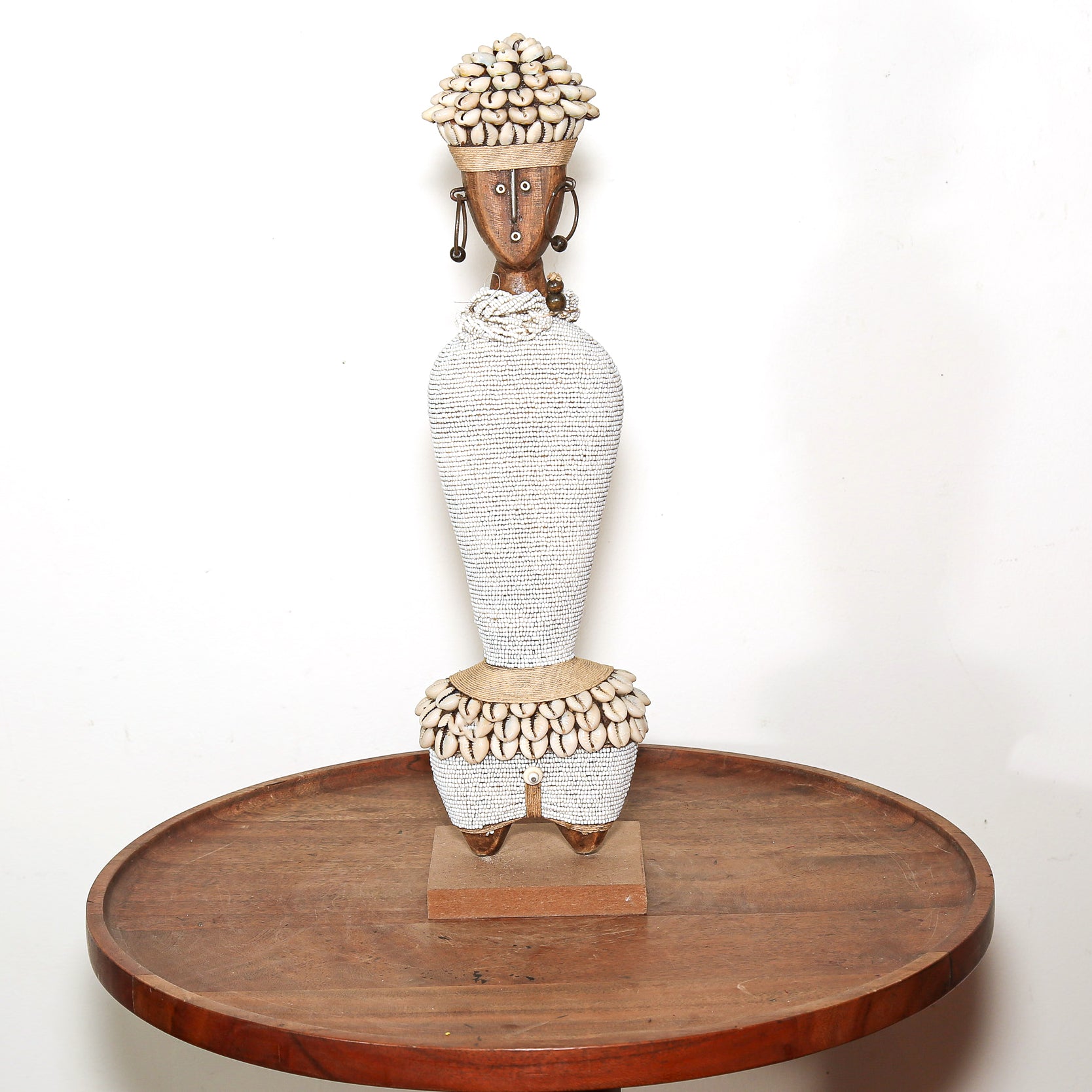 A tall African-style figurative sculpture of a woman with a textured white body, natural wood face and base, and beige shell-like hair and waist details, displayed on a round wooden table against a white wall.