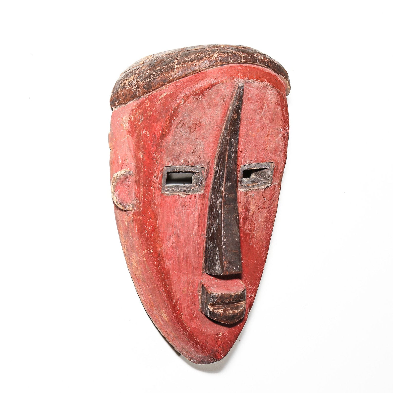 A hand-carved wooden mask with a red-painted face and dark brown painted details, including the nose, mouth, and hair-like top feature, shown against a plain white background.