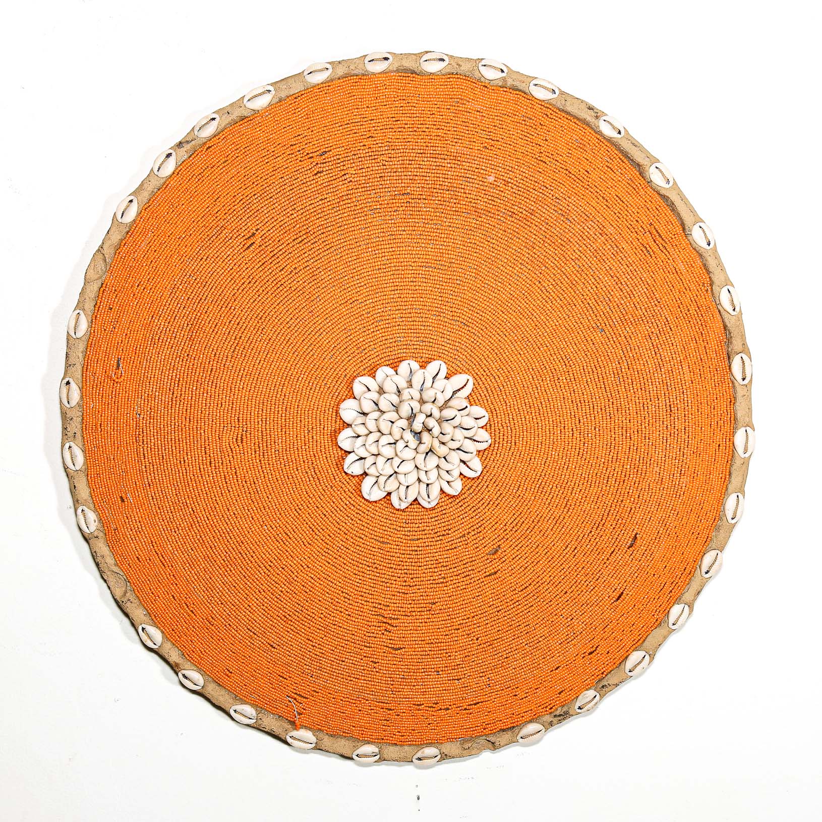 Round orange beaded wall shield decor with a central white flower design made from cowrie shells hanging on a white wall.