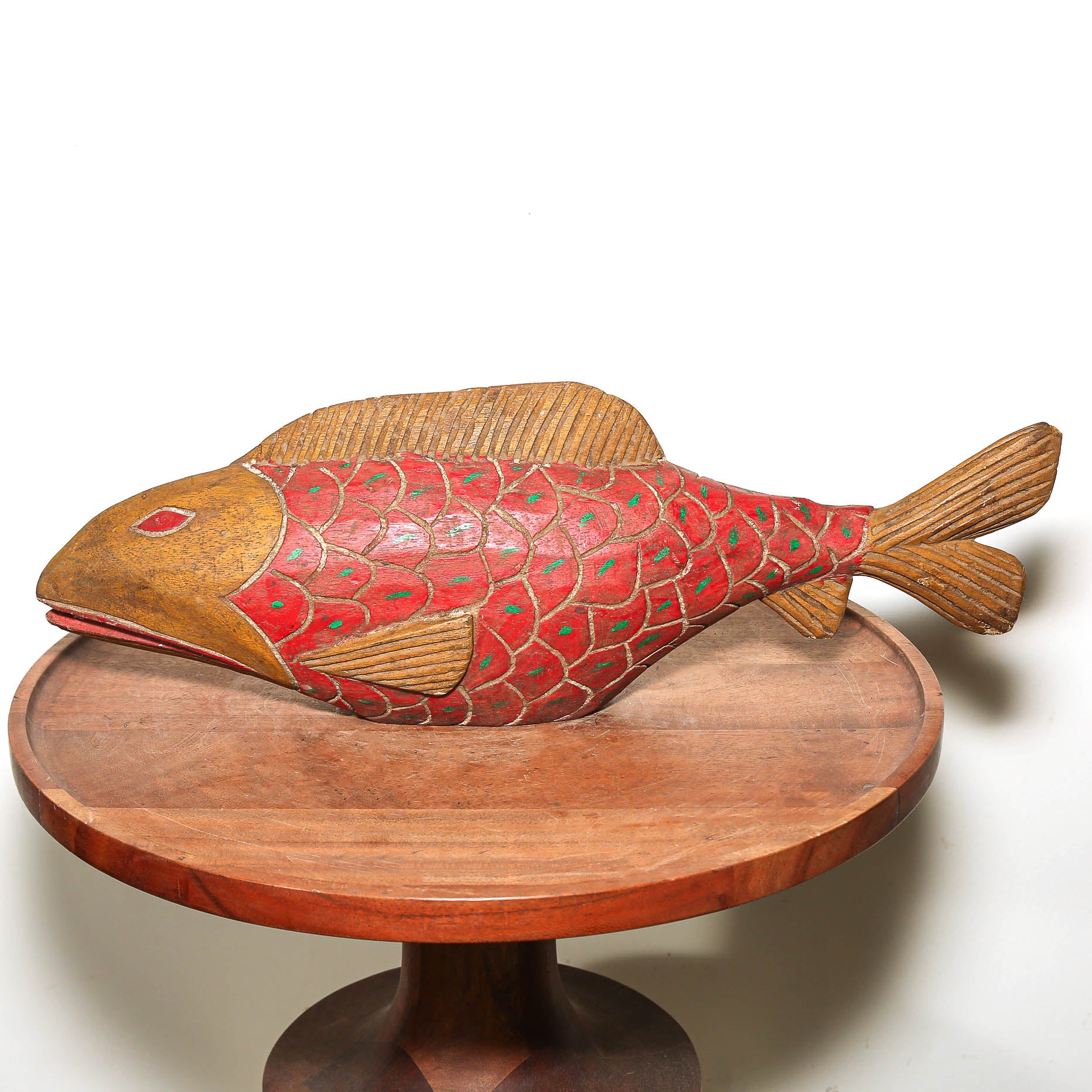 A hand-carved wooden fish sculpture with a red and green patterned body and natural brown wood edges, displayed on a round wooden pedestal against a white background.