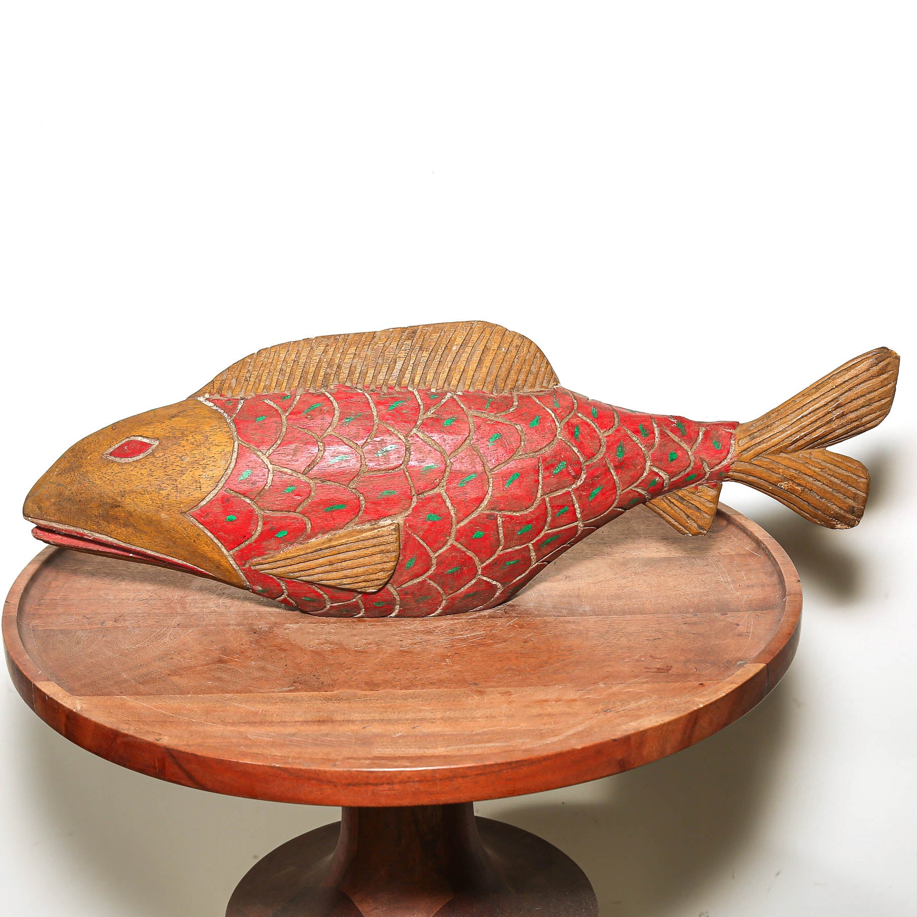 A hand-carved wooden fish sculpture with a red and green patterned body and natural wood fins, displayed on a round wooden pedestal against a white background.