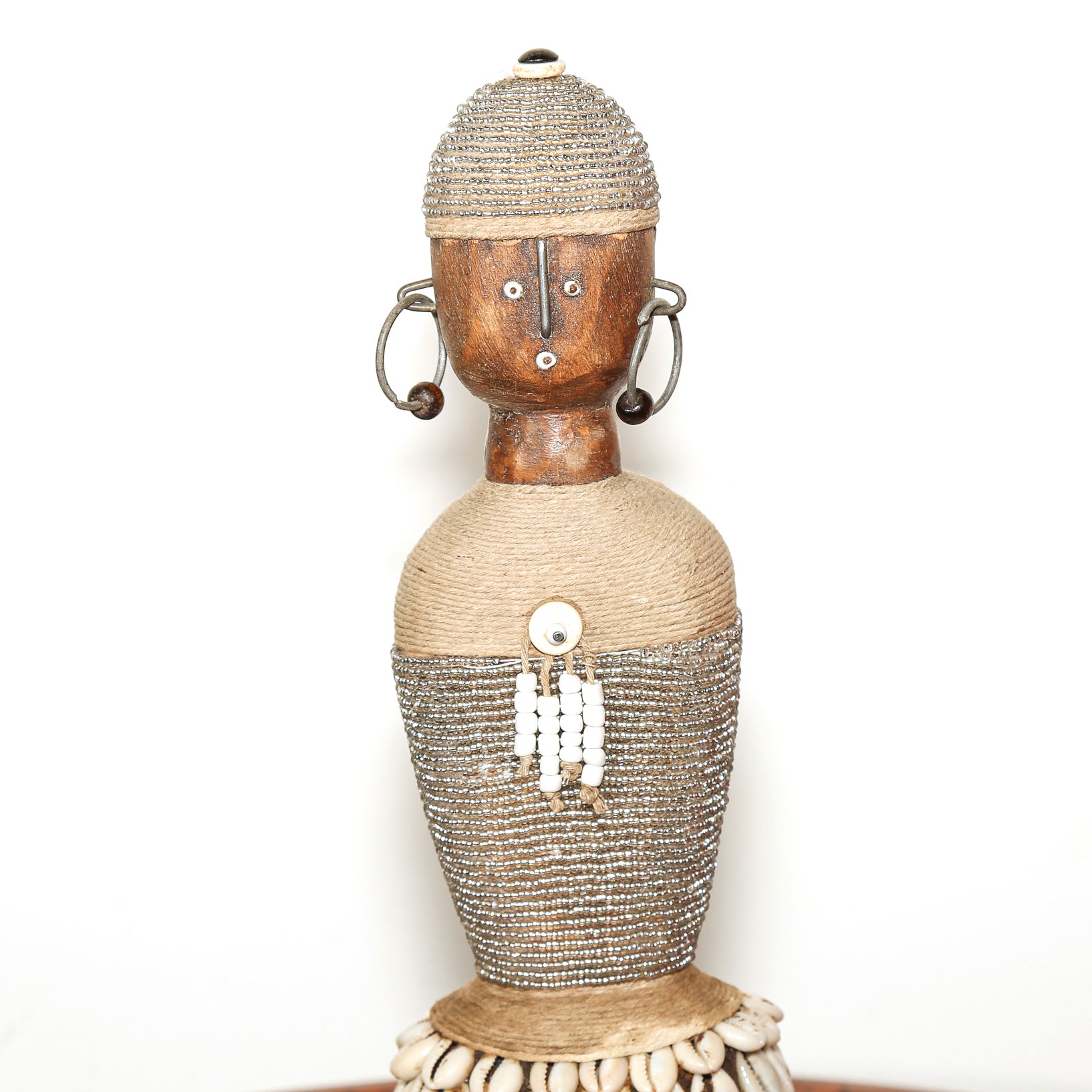 A handmade African-style wooden doll figurine with a carved brown face, woven beige and metallic silver beadwork, and silver hoop earrings, set against a plain white background.